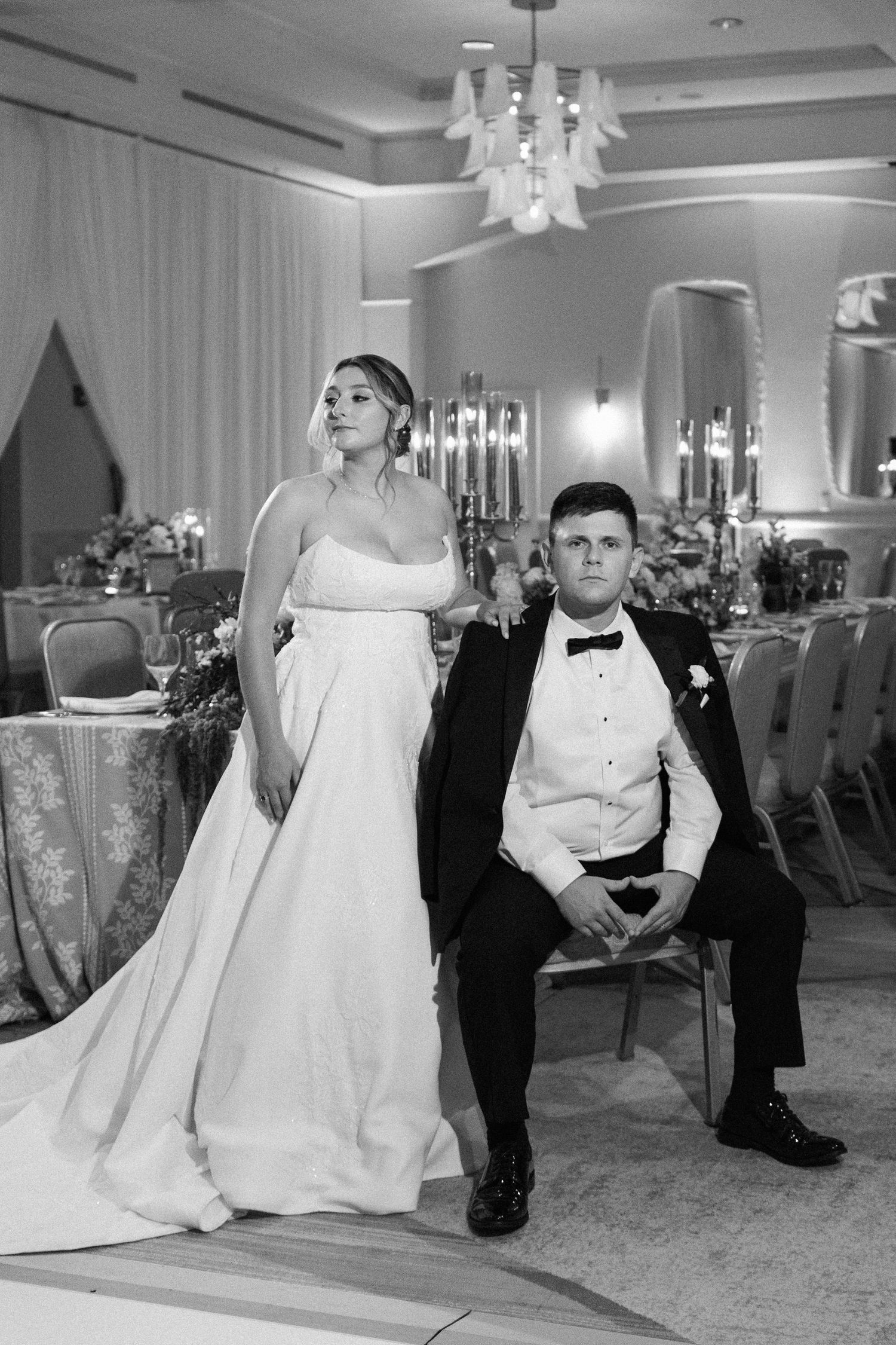Black and white photo of a bride in wedding dress and veil sitting in a sunlit room, viewed through a dark hallway.
