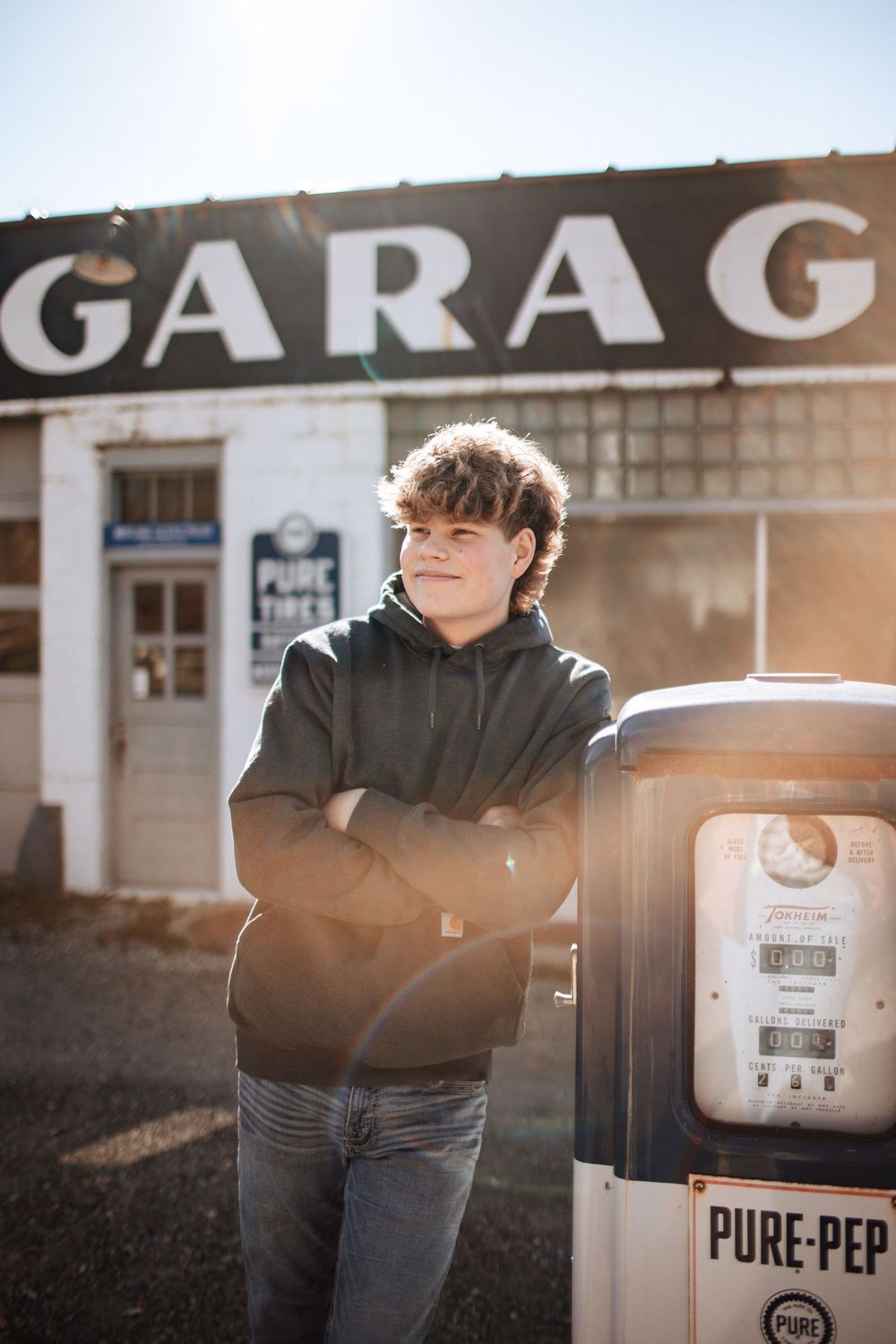 Teen boy in gray hoodie leans on vintage Pure-Pep gas pump outside retro garage on sunny day.