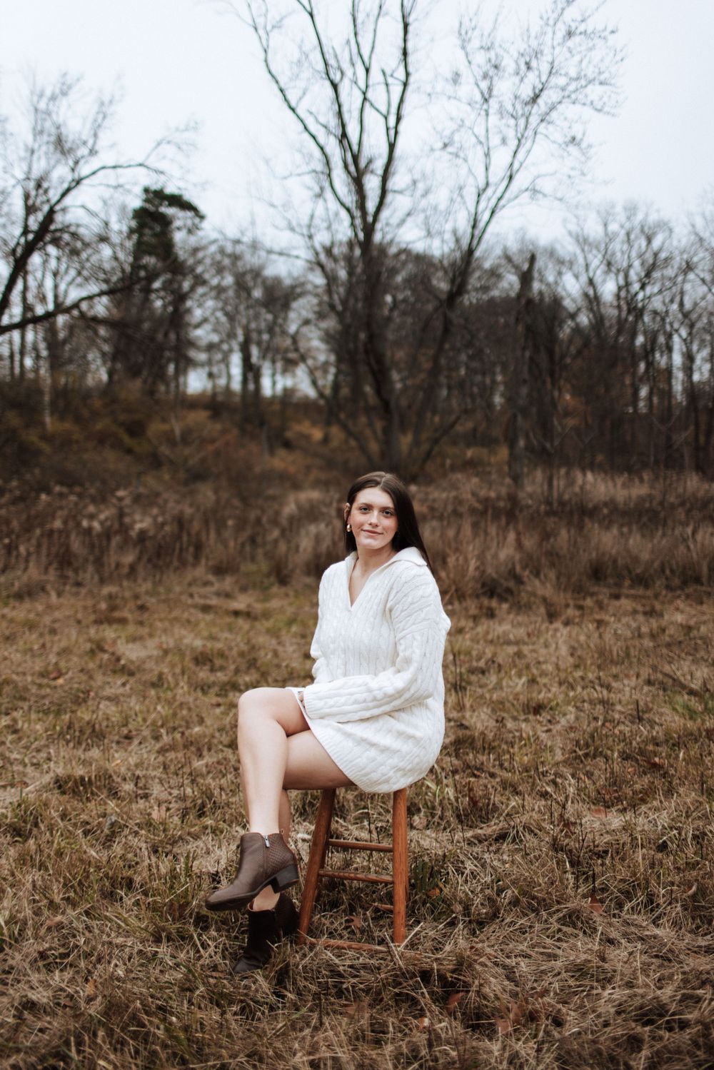 Young woman in white knit dress sitting on a stool in a bare winter field.
