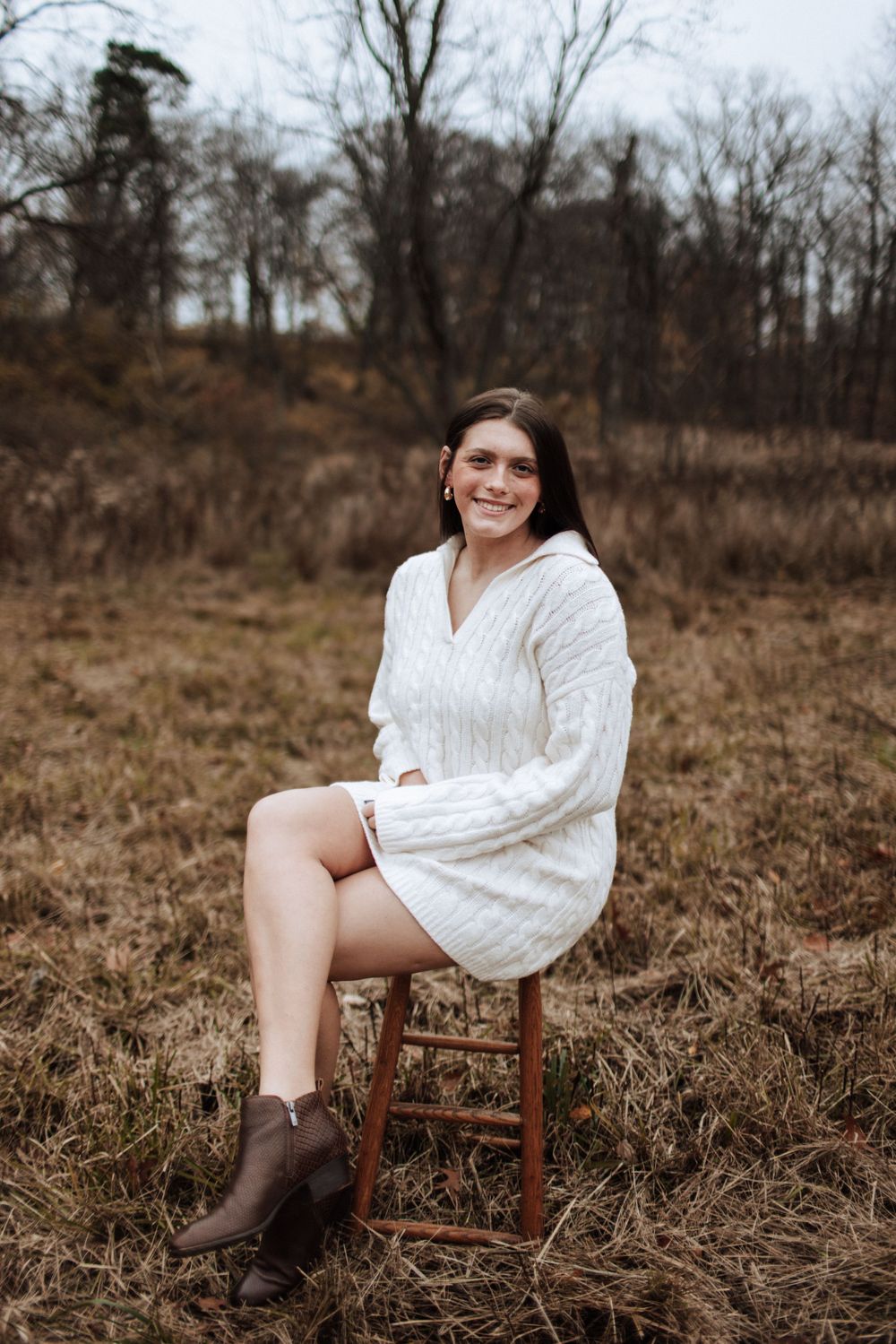 Brunette girl in white cable knit sweater dress sits on stool in winter field.