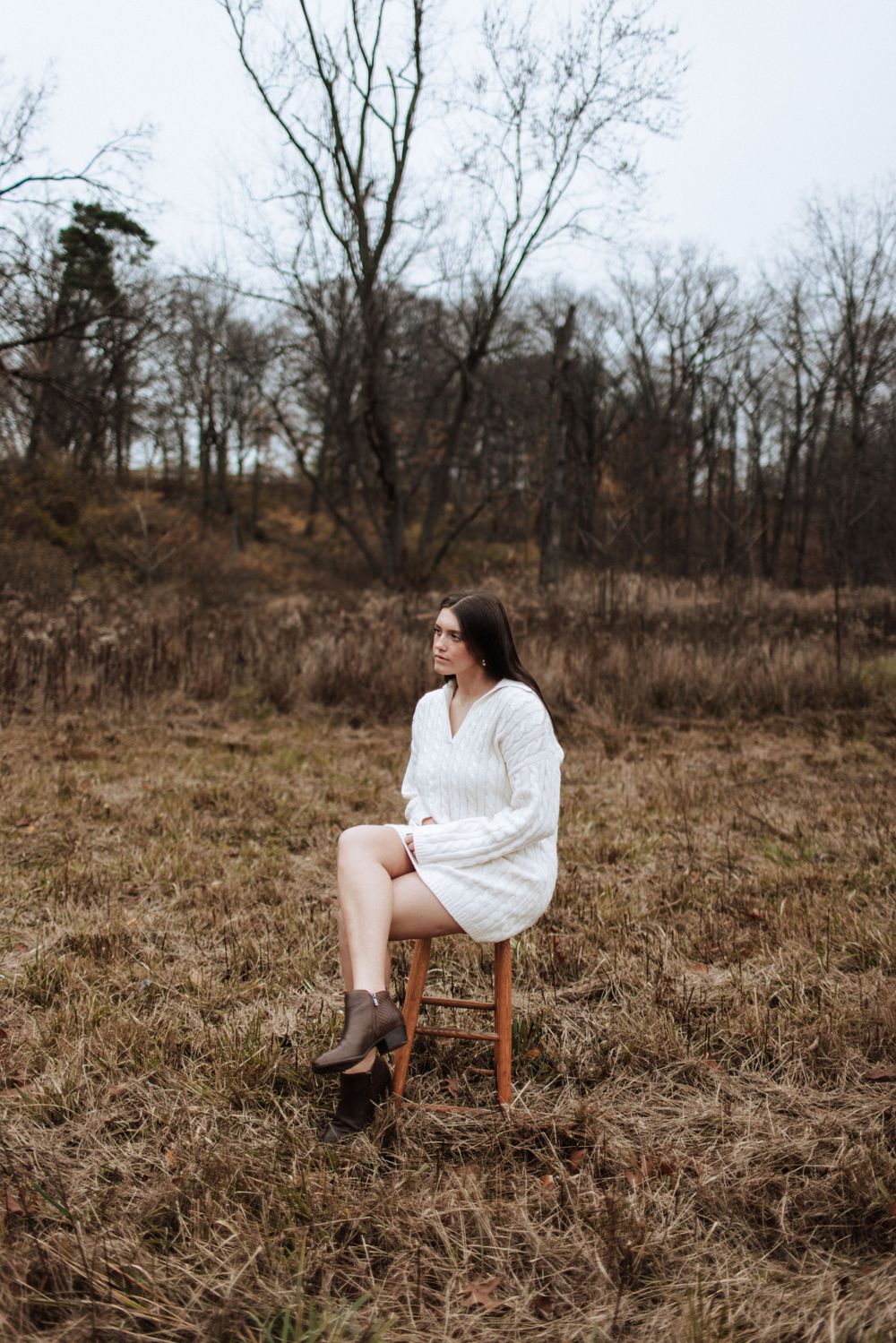 Young woman in white knit sweater dress sits on stool in barren winter field.