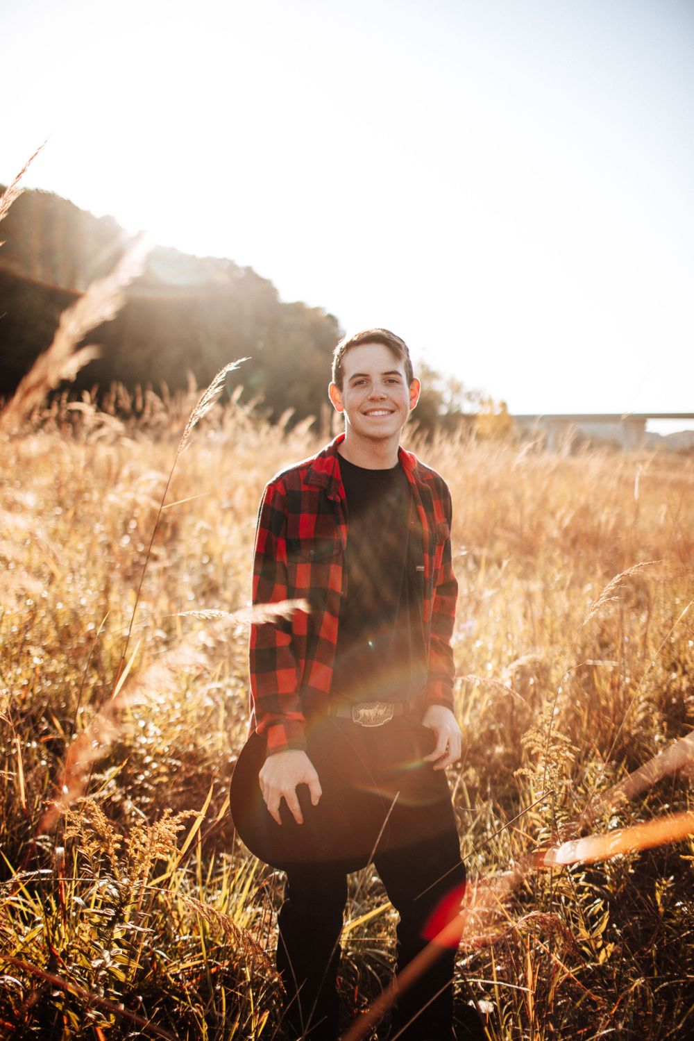 Smiling young man in red plaid shirt standing in a sunlit golden autumn field.