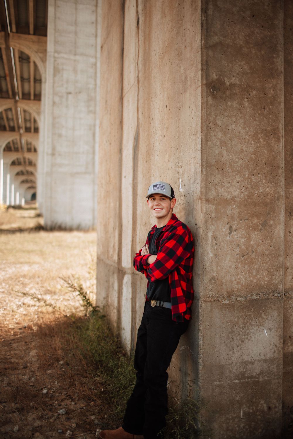 Smiling young man in red plaid shirt leans against concrete bridge pillar.