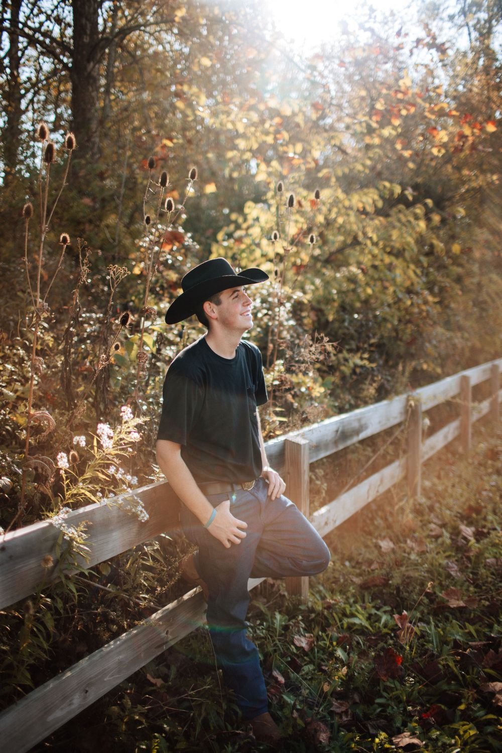 Young man in black cowboy hat leaning on a white fence in a sunlit autumn garden.