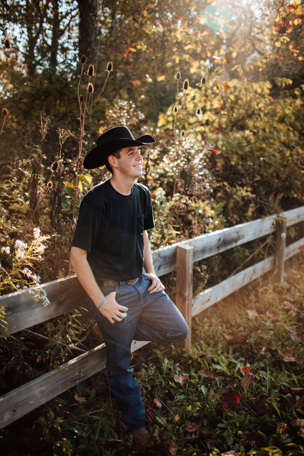 Young man in black cowboy hat leans on wooden fence in autumn garden.