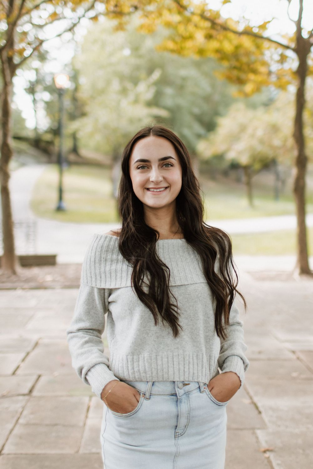 Young woman with long dark hair smiling in gray off-shoulder sweater and jeans in autumn park setting.