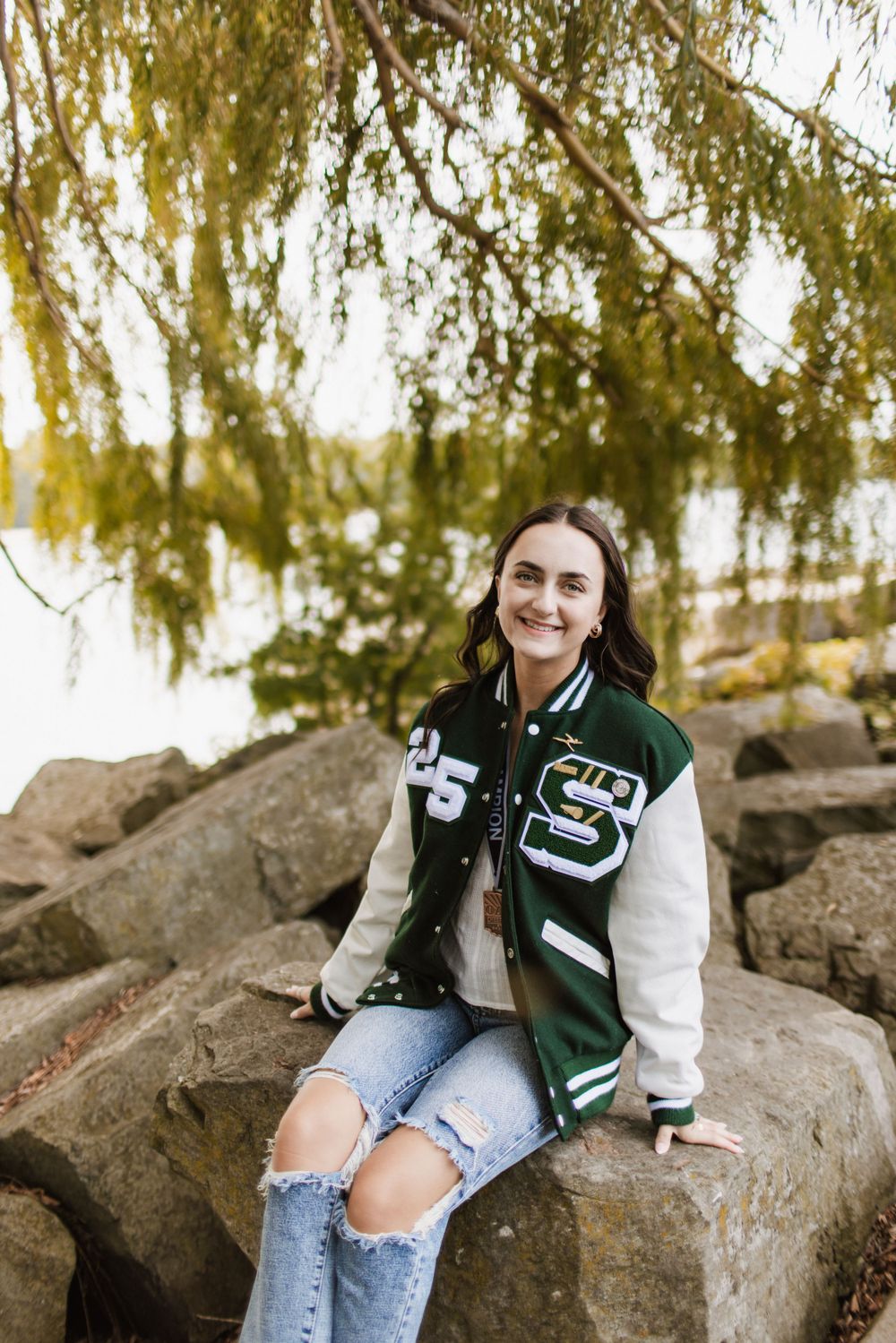 Smiling teen girl in green varsity jacket sitting on rocks under weeping willow tree outdoors.