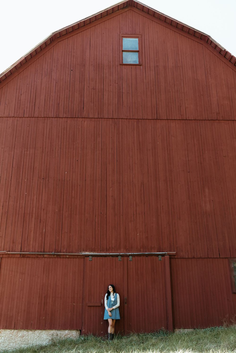 Young woman leaning against a large red barn door in a rural farm setting.