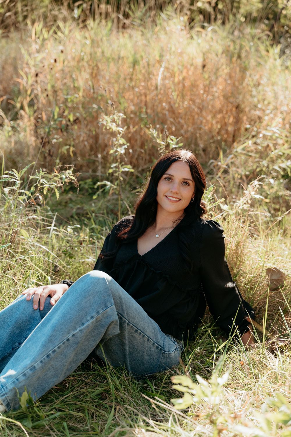 Woman in black blouse and jeans sitting in a sunny field with tall golden grasses in the background.