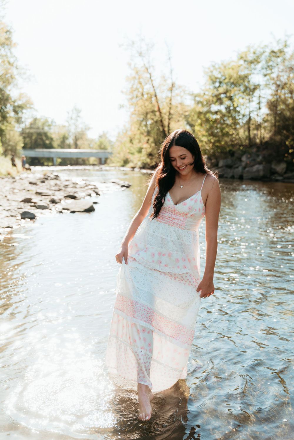 A smiling woman in a white maxi dress wades through a shallow river on a sunny day.