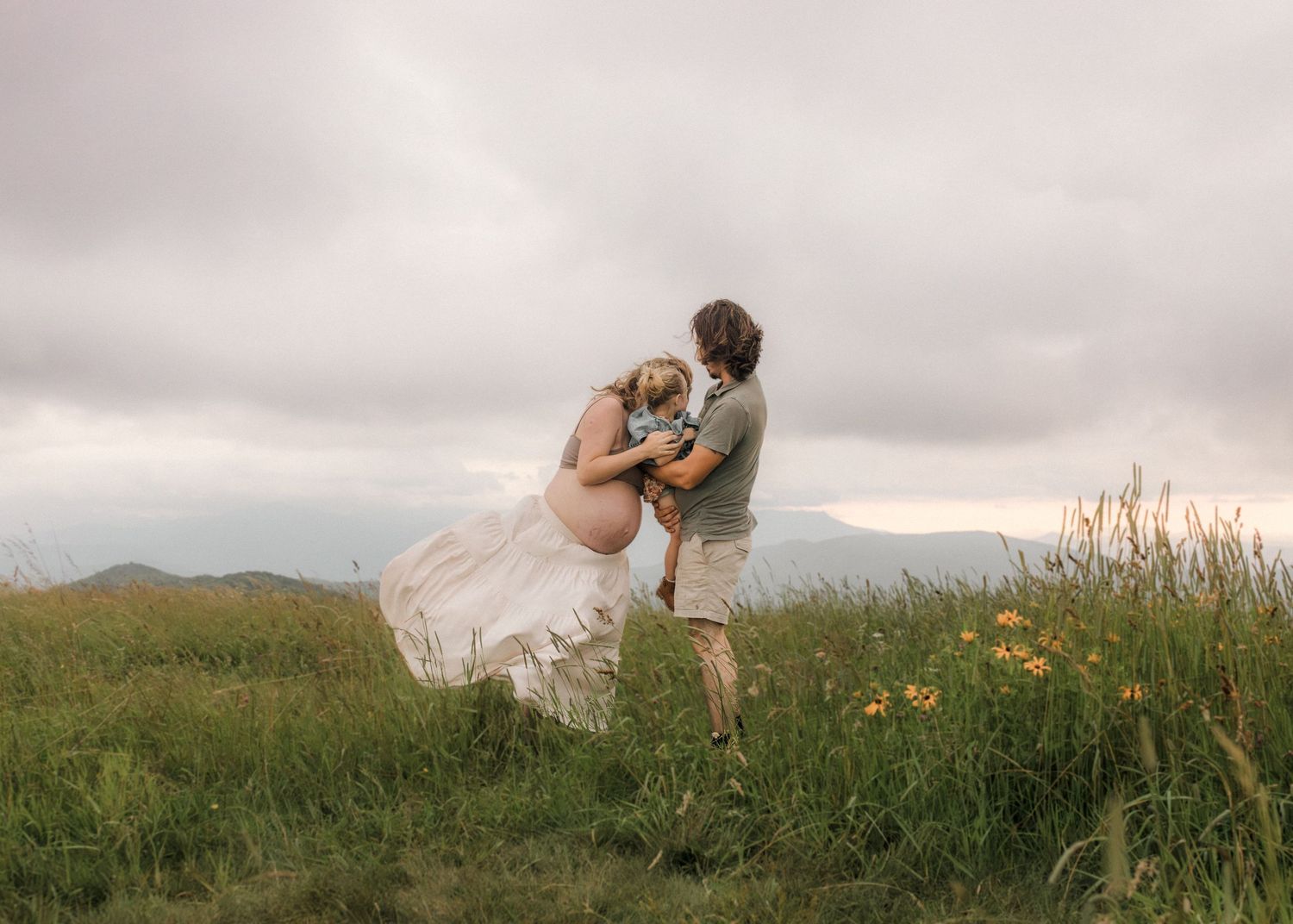 Pregnant couple embracing in a wildflower meadow with mountain views and dramatic cloudy sky.