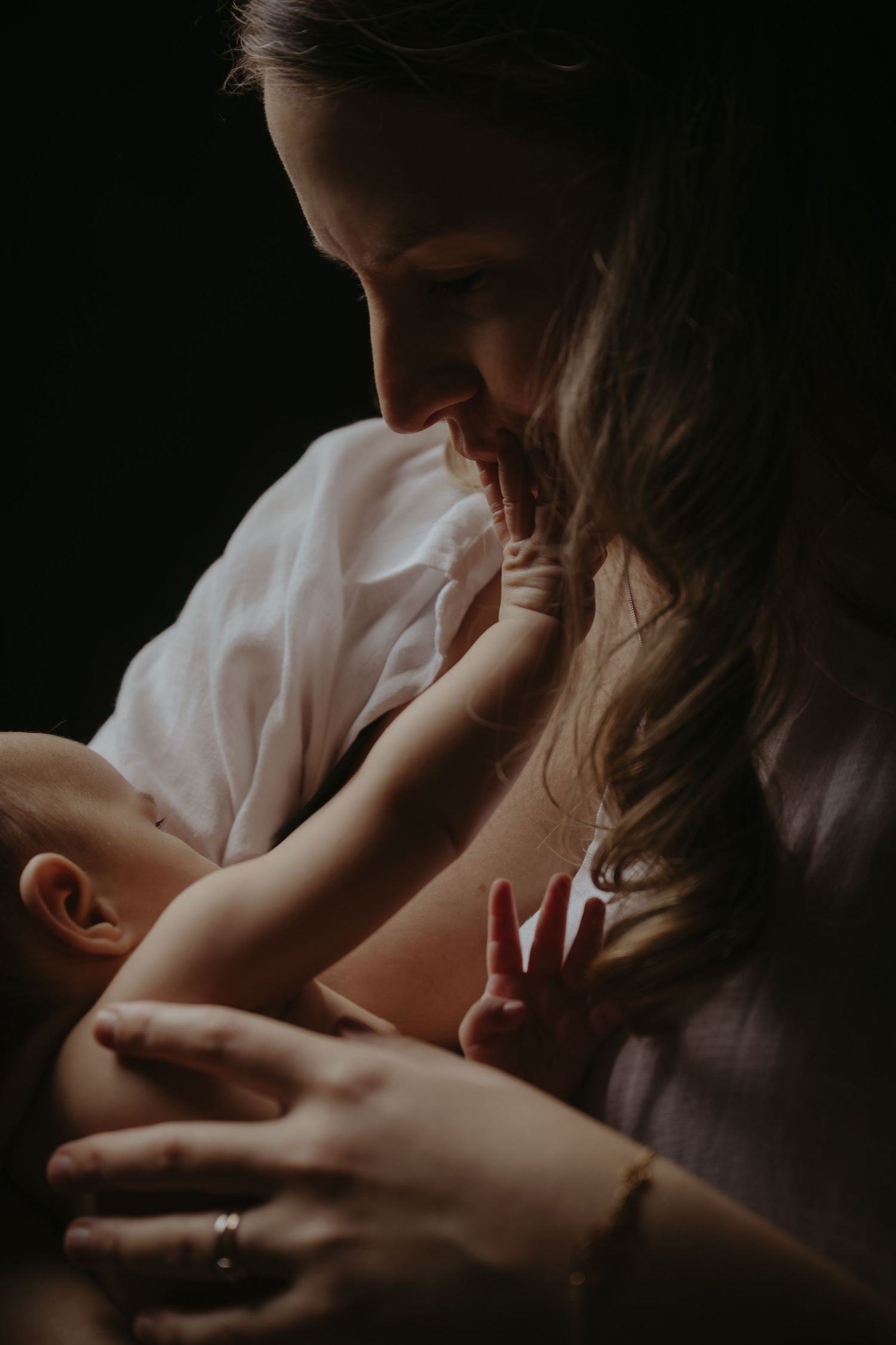 A mother tenderly holds her newborn baby in a softly lit, dark background portrait photo.