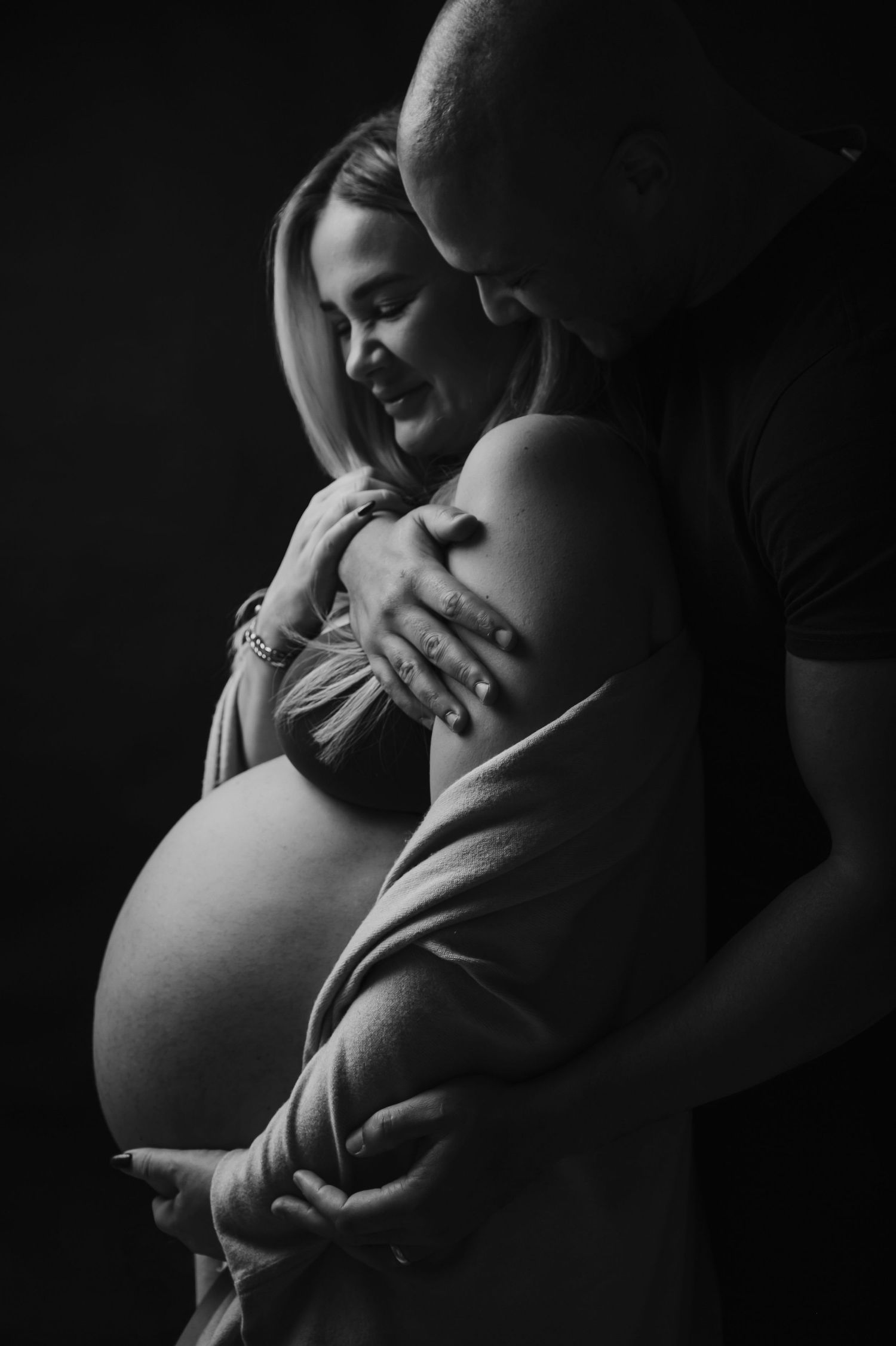 Black and white maternity photo of a couple embracing, man kissing woman's head while cradling her pregnant belly.