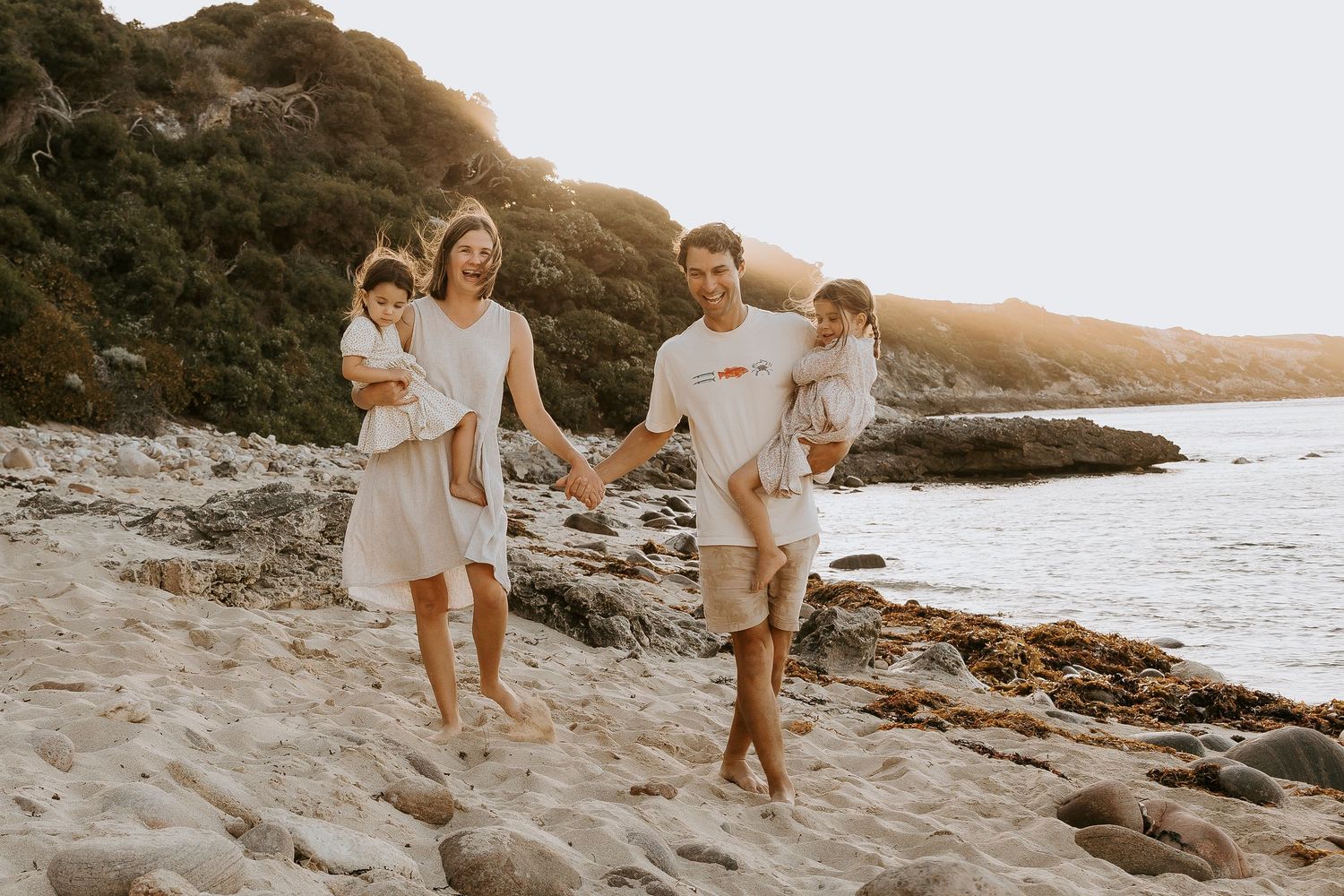 Family of four smiling together on rocky beach at golden sunset with ocean backdrop.