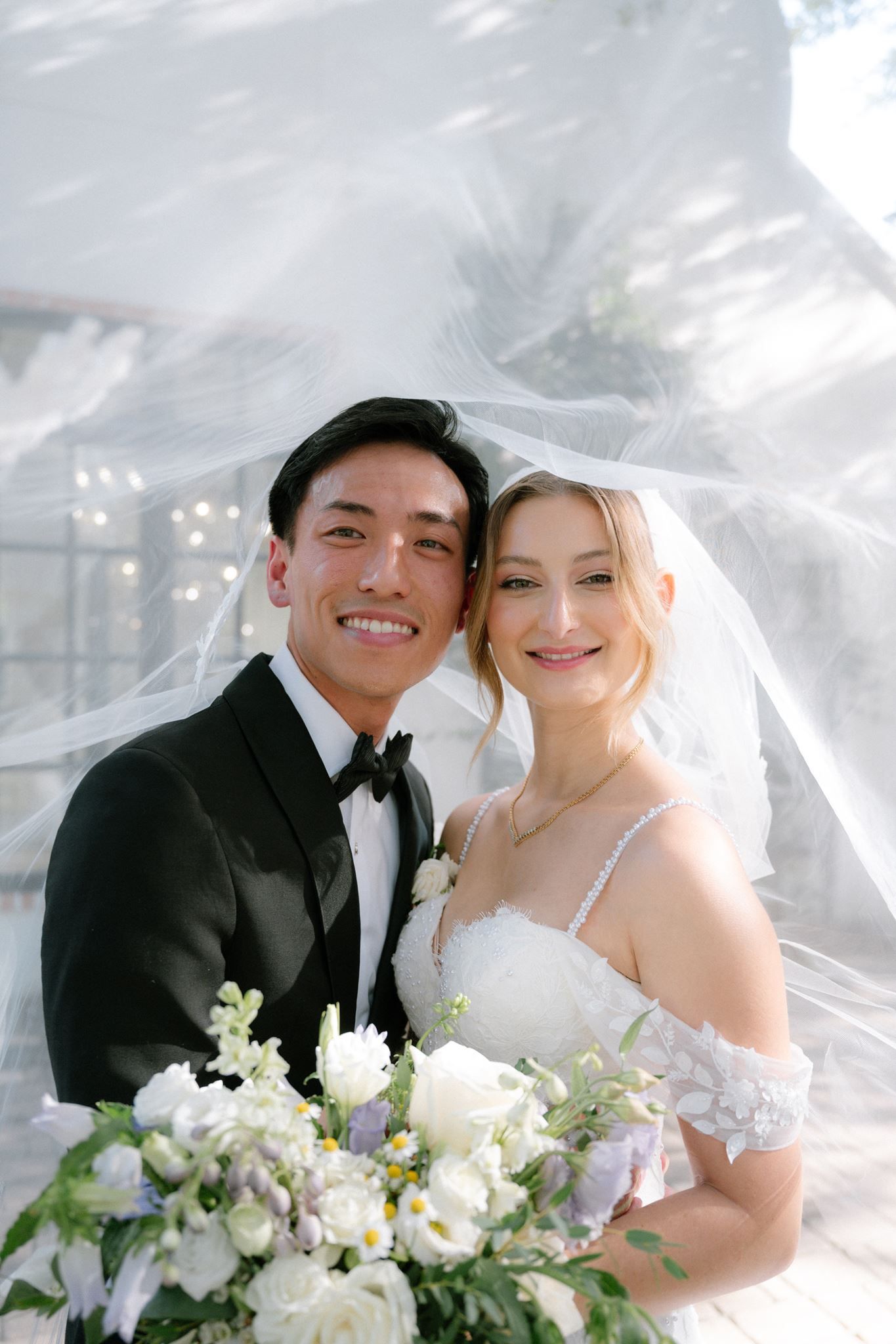 A smiling bride and groom pose together under a flowing white veil, bride holding a white floral bouquet.