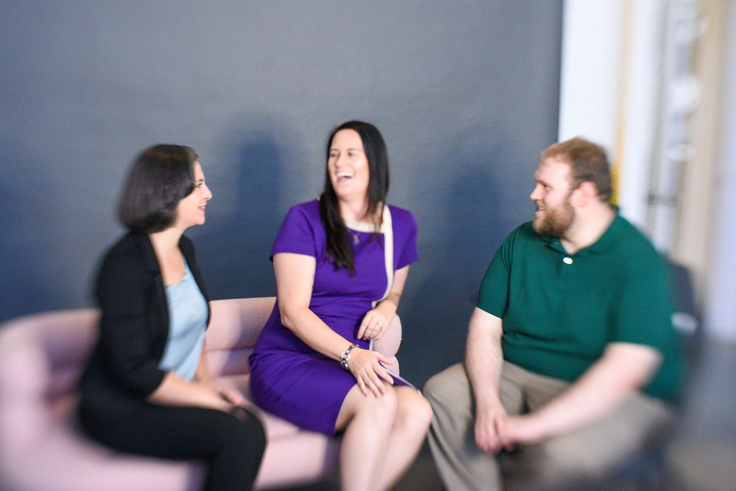Three colleagues laughing and having a friendly conversation while seated together in a modern office lounge area.