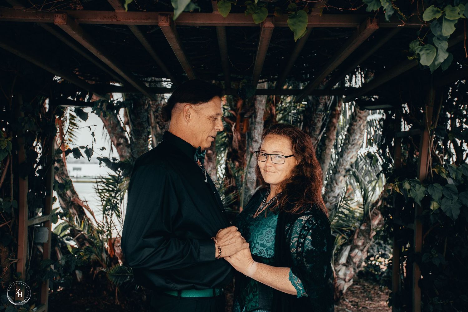 Couple dancing together in a lush greenhouse setting, man in black suit and woman in teal dress.