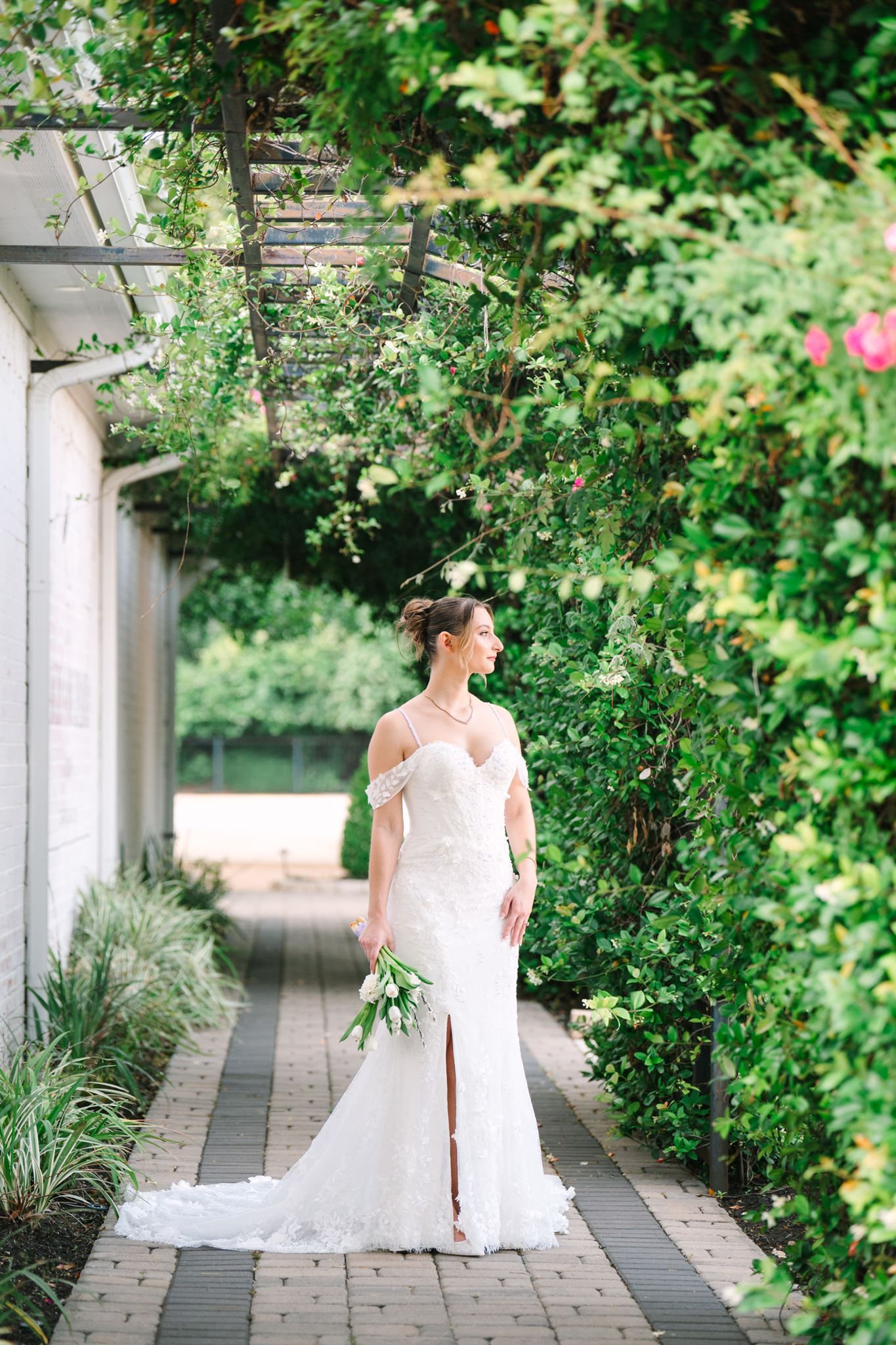 Bride in white off-shoulder gown holding bouquet stands under lush green pergola on brick pathway.