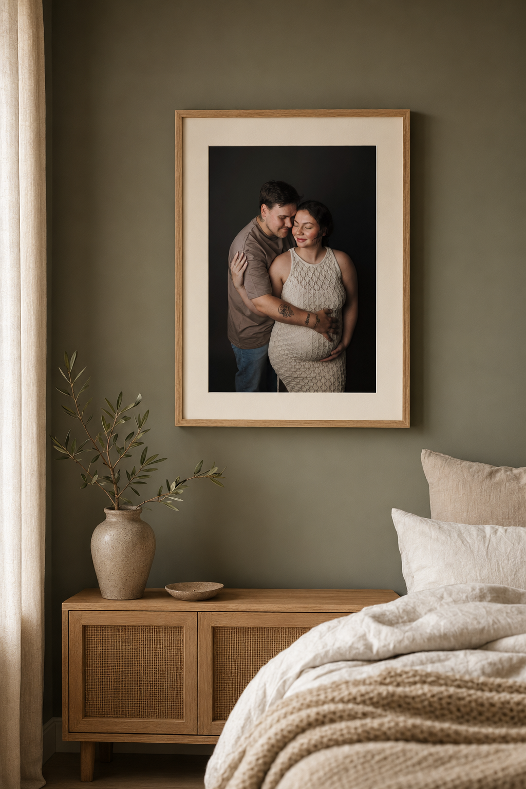Framed maternity photo of a couple displayed on a sage green bedroom wall above a wooden dresser with a ceramic vase.
