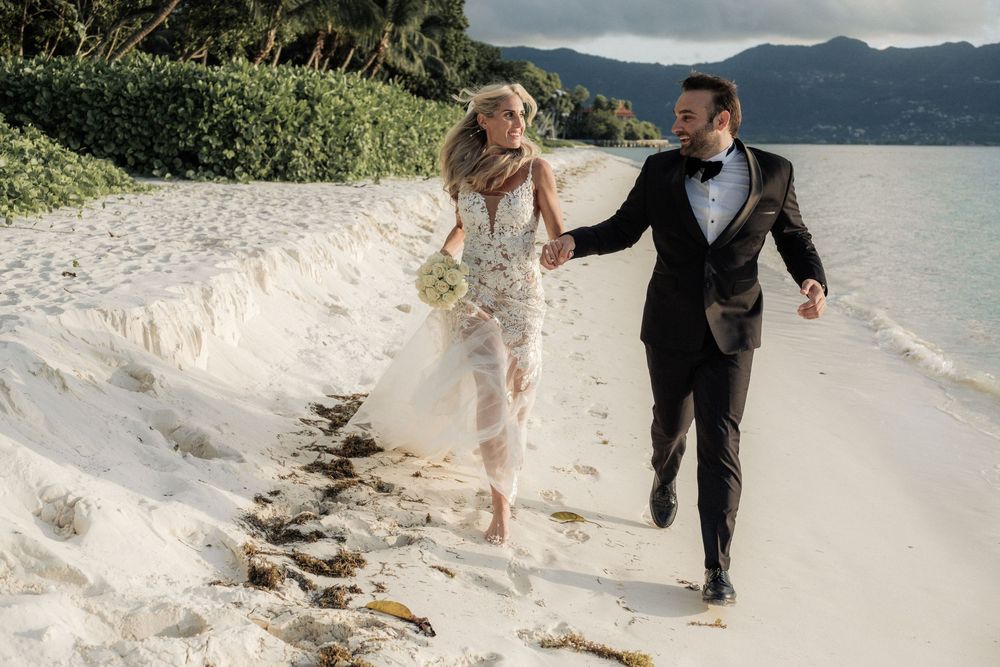 Couple in wedding attire embracing on a beach at sunset with mountains and ocean in the background.