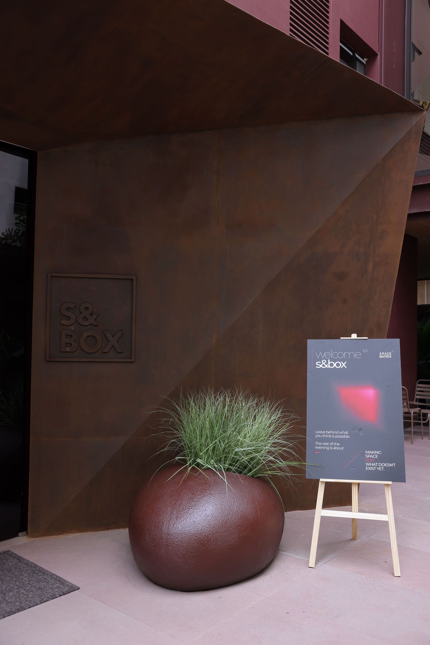 S&Box building entrance with rustic metal facade, large round planter with grass, and welcome signage on wooden easel.