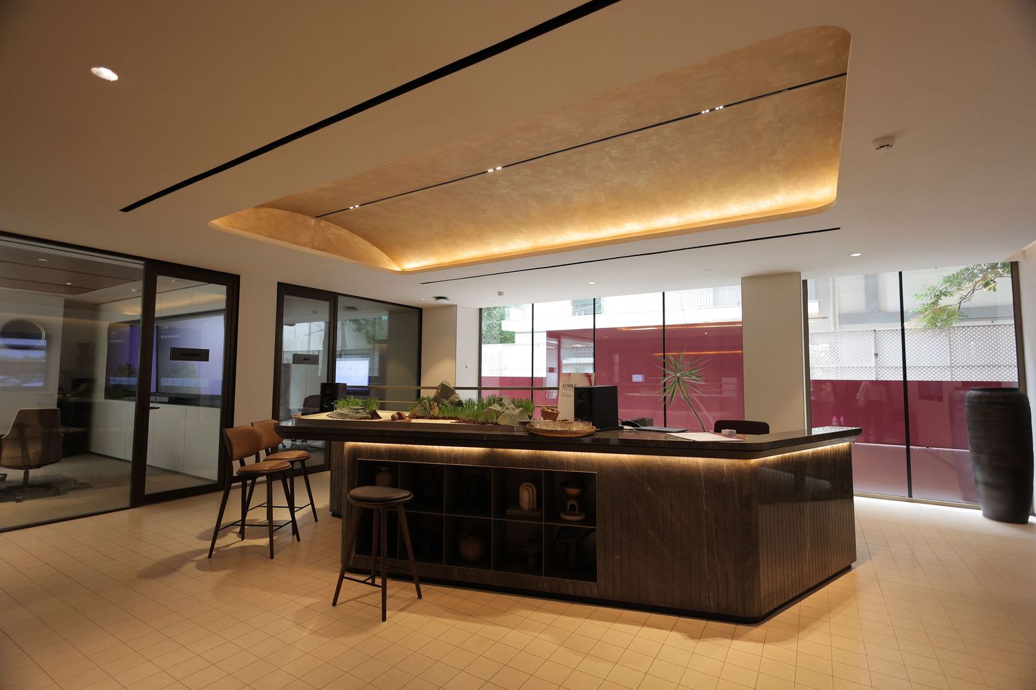 Modern office lobby with dark wood reception desk, warm ceiling lighting, bar stools, and floor-to-ceiling windows.