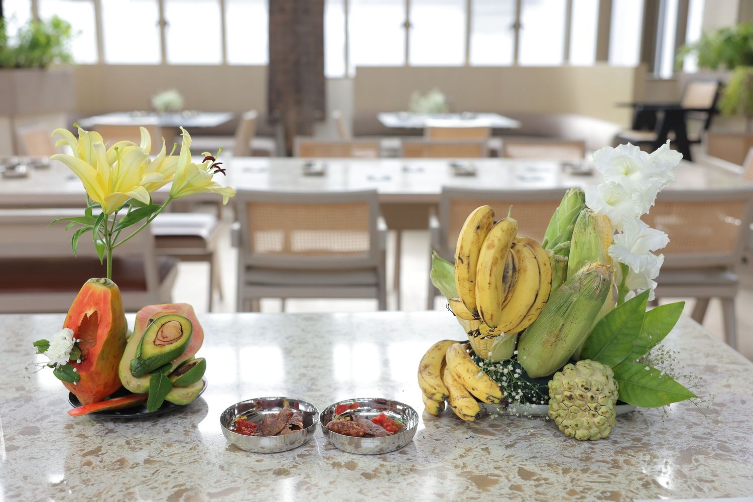Tropical fruit display with bananas, papaya, and flowers on a marble table in a bright restaurant dining room.