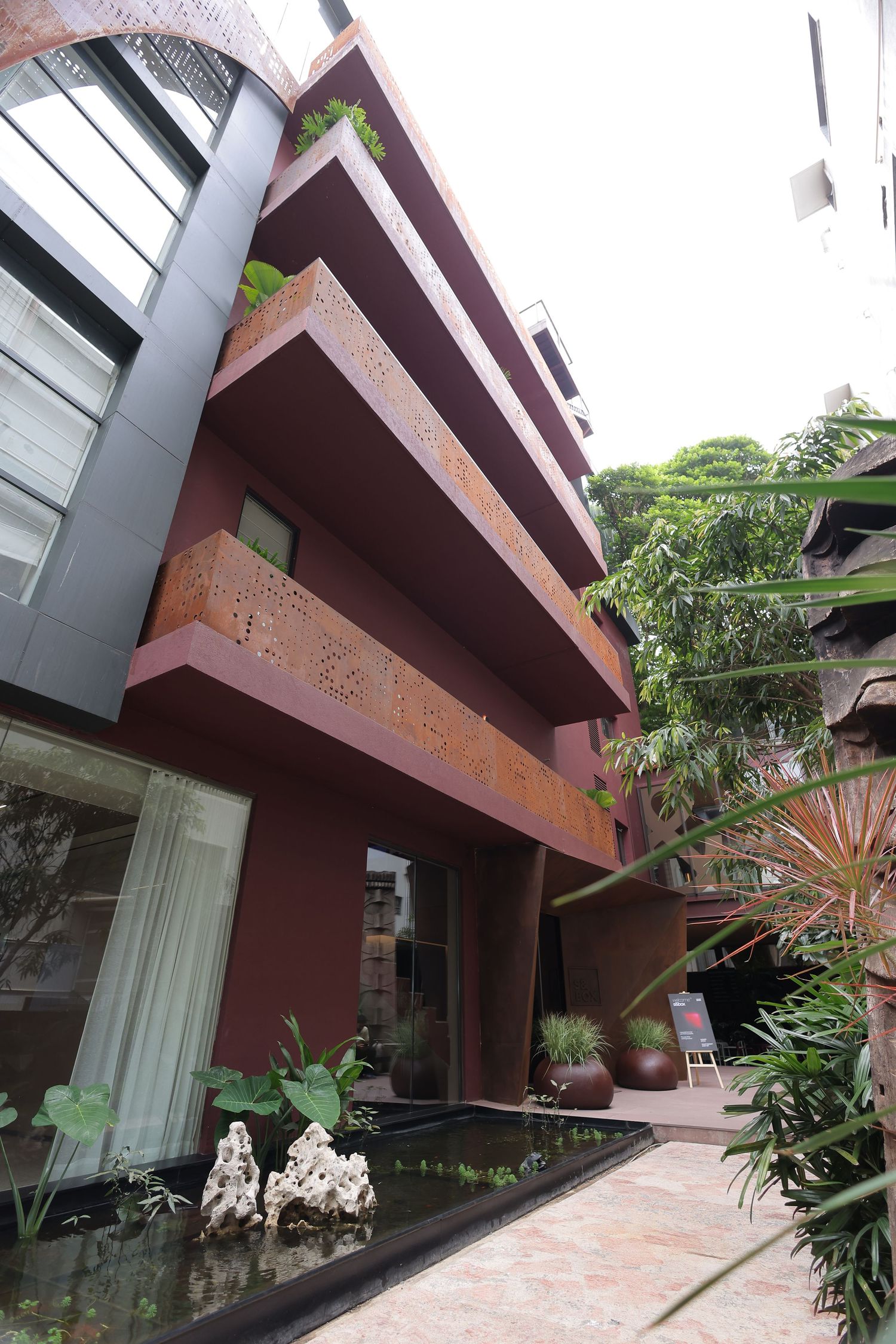 Modern red building exterior with lush greenery, decorative balconies, and a serene water feature with white rocks.