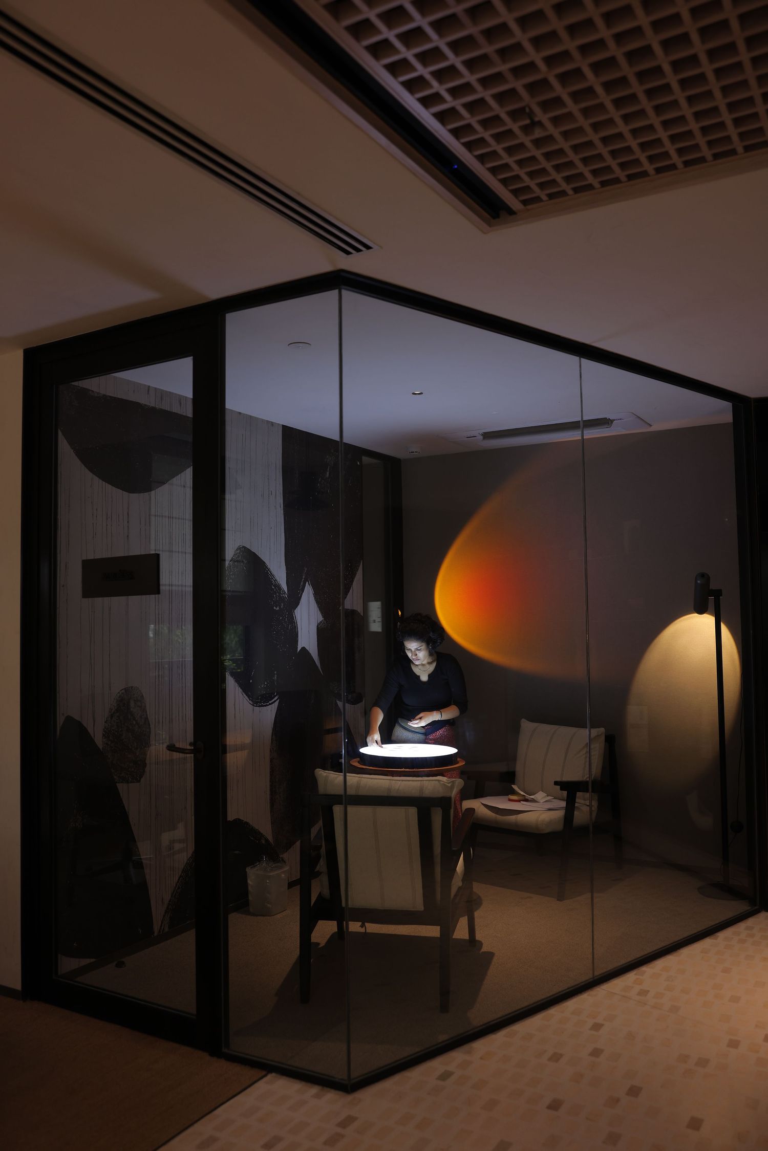 Person working at illuminated table inside modern glass-walled room with warm ambient lighting and contemporary decor.