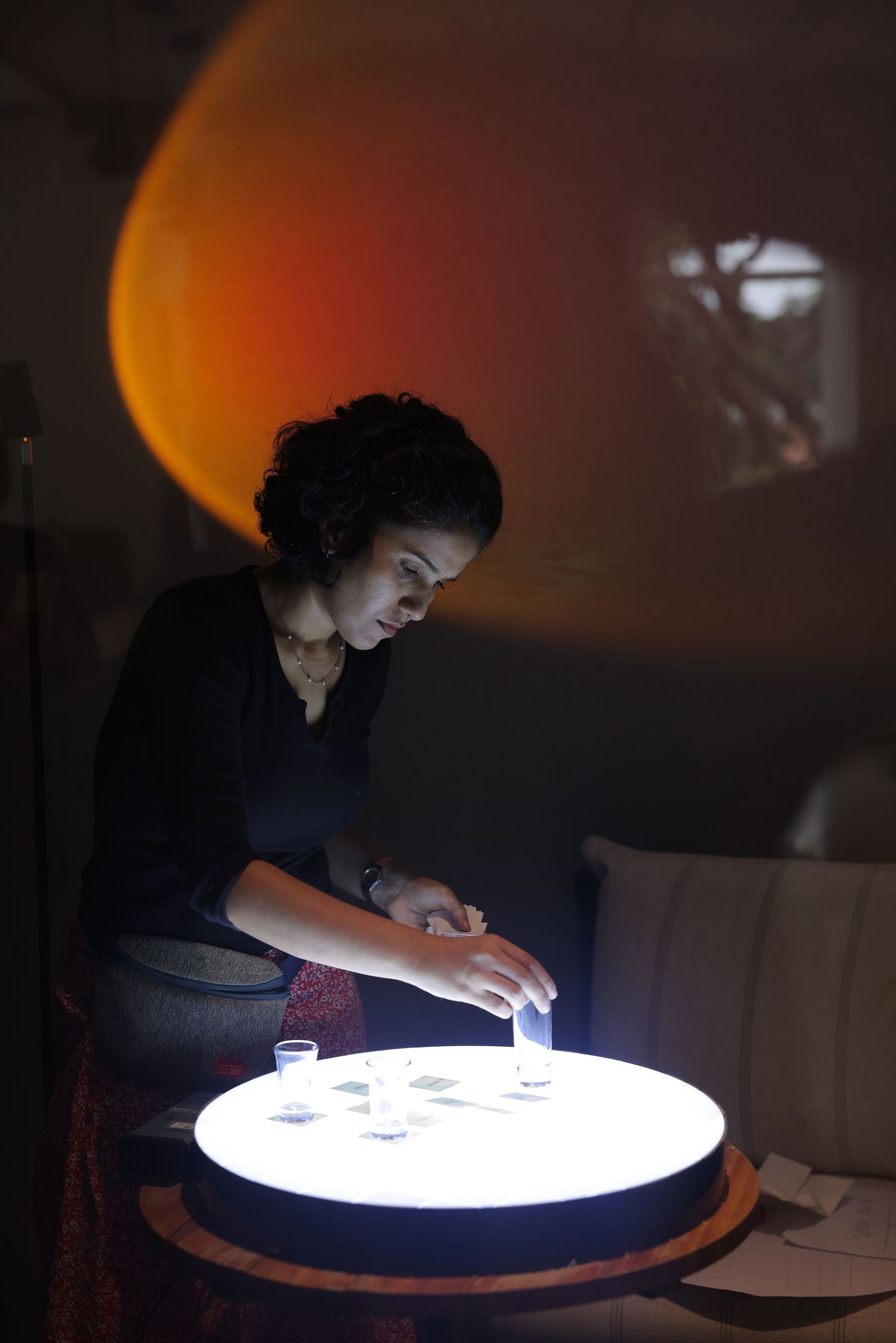 Woman examining glowing illuminated table with small glasses in a dimly lit room with orange ambient lighting.