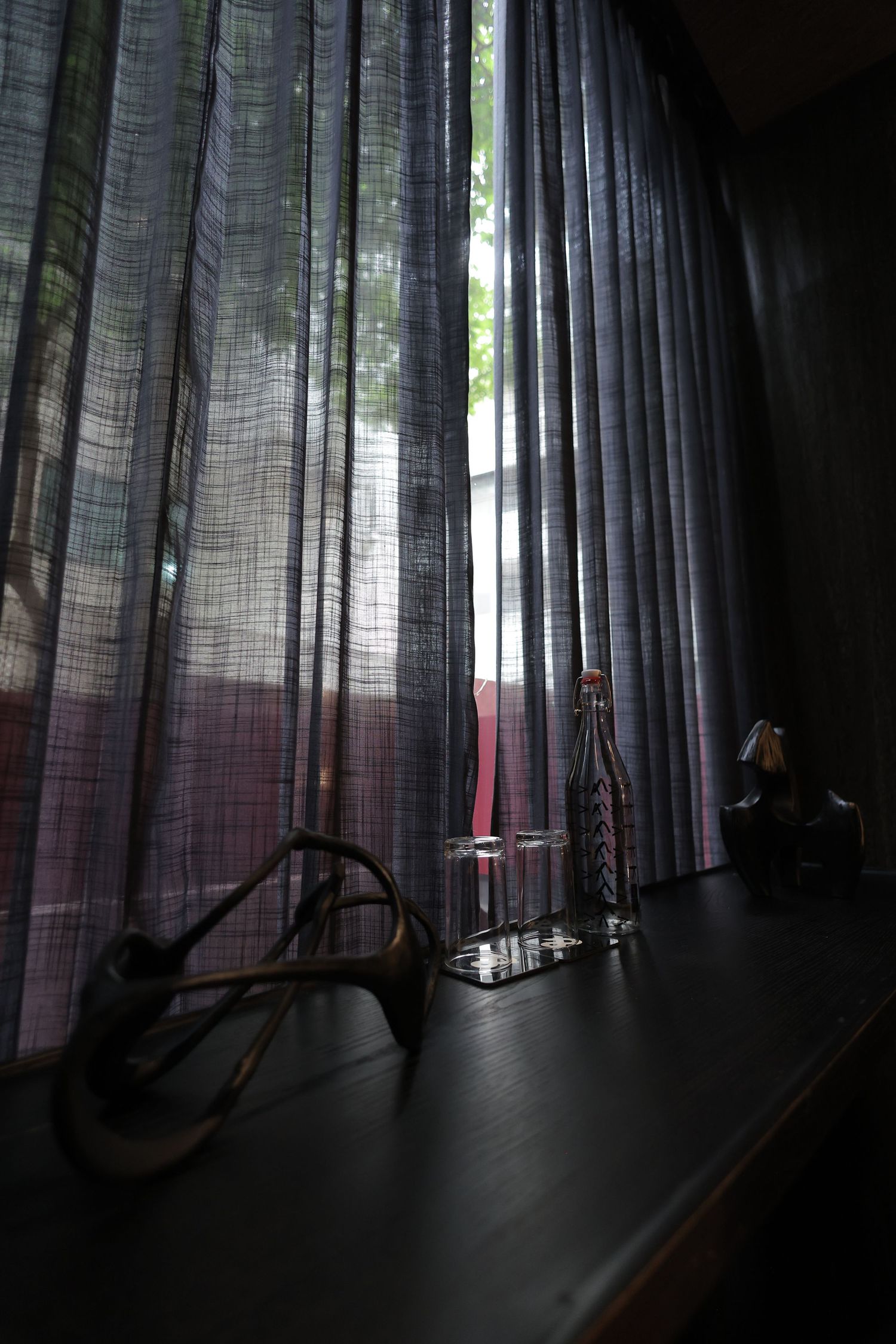Sheer curtains filter natural light over a dark shelf displaying decorative objects including a bottle and figurines.