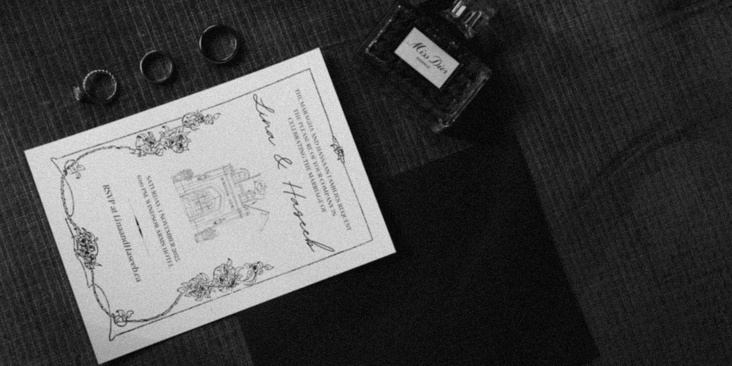 Black and white flat lay of wedding invitation, rings, ink bottle, and envelope on textured fabric.