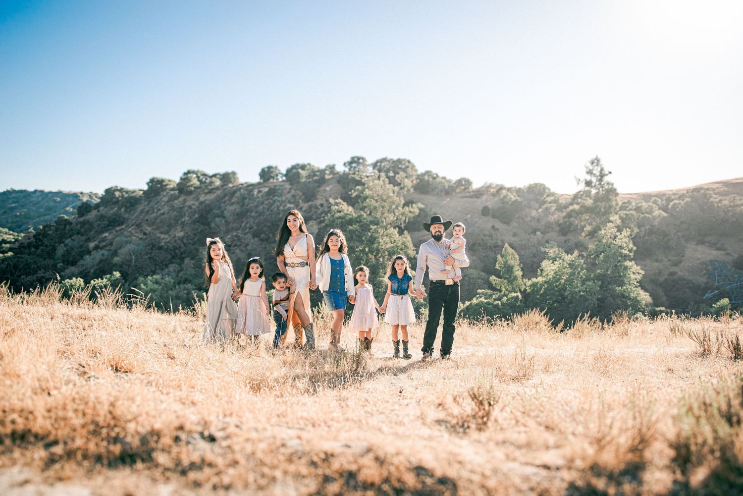 Large family posing together on a sunny golden hillside with lush green trees in the background.