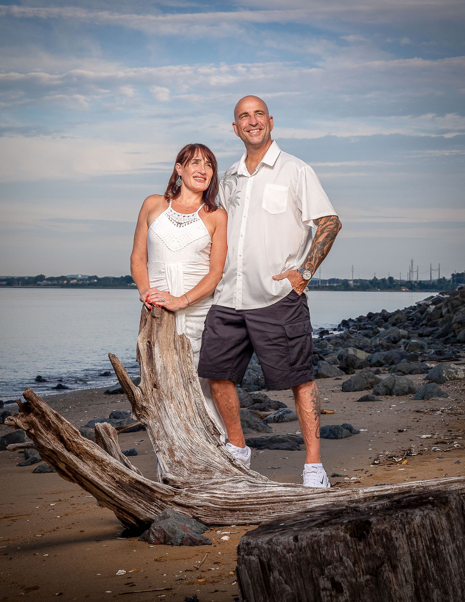 Couple posing on driftwood at a rocky beach shoreline during golden hour with calm water in the background.