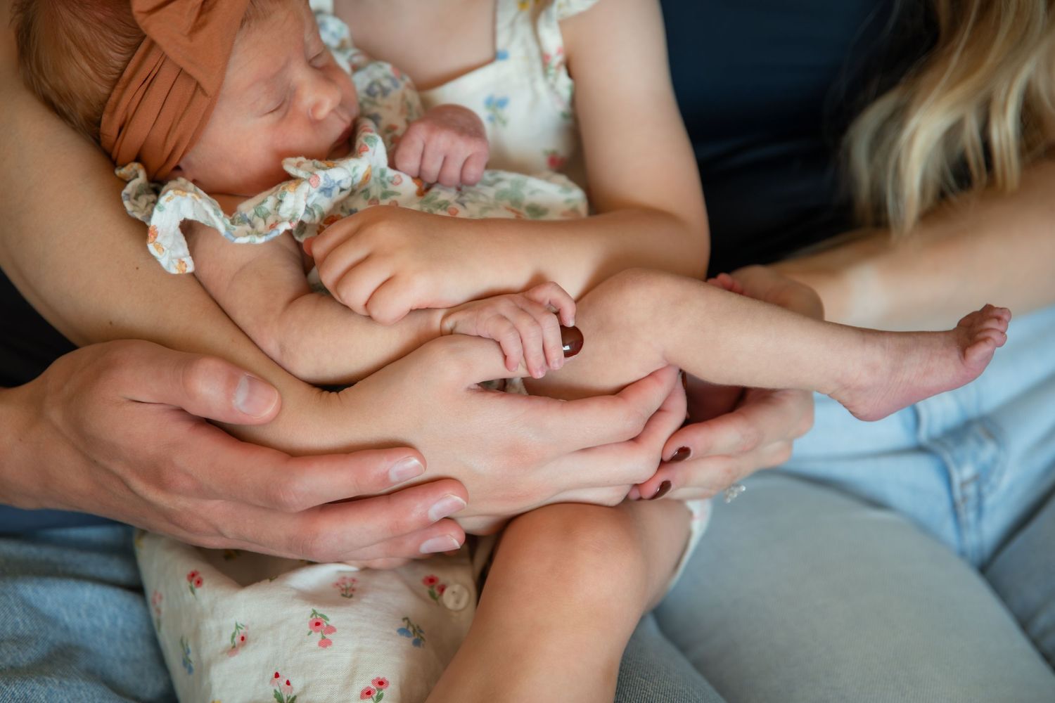 Young couple sitting on couch kissing while holding toddler and newborn baby wrapped in orange bow headband.