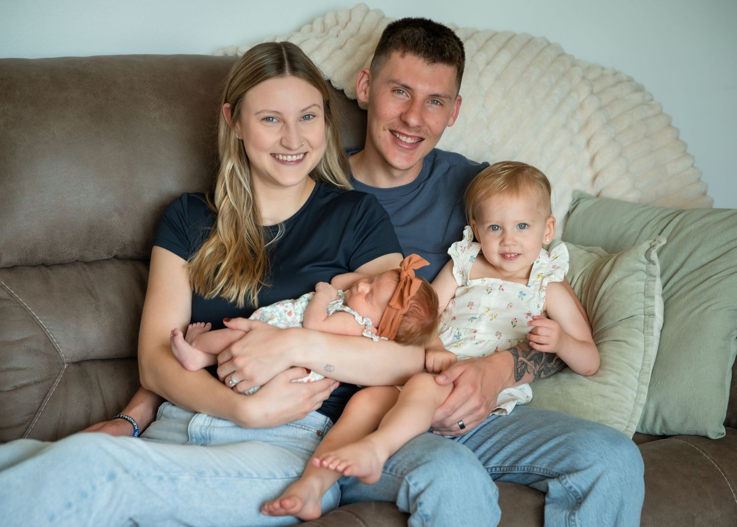Young family of four sitting on a couch, parents smiling while holding a newborn and a toddler girl.