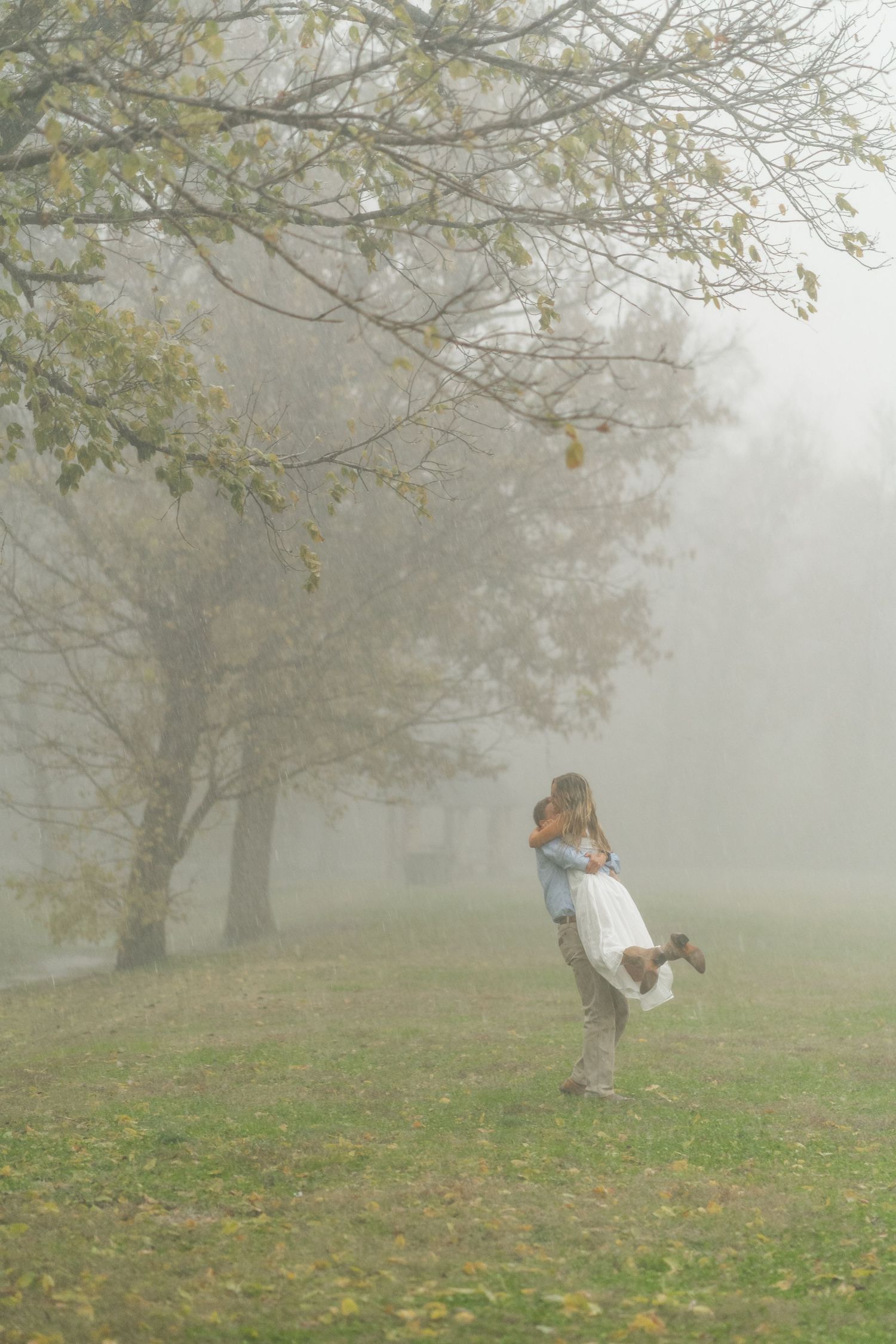 Couple sharing a romantic moment in a misty foggy park, woman in white dress and man in blue shirt holding hands.
