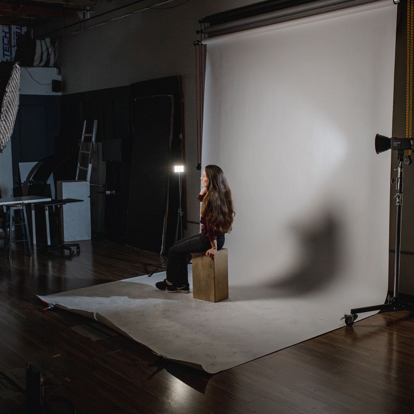 Woman in red shirt and black pants sits on a stool in a photography studio with white backdrop and lighting.