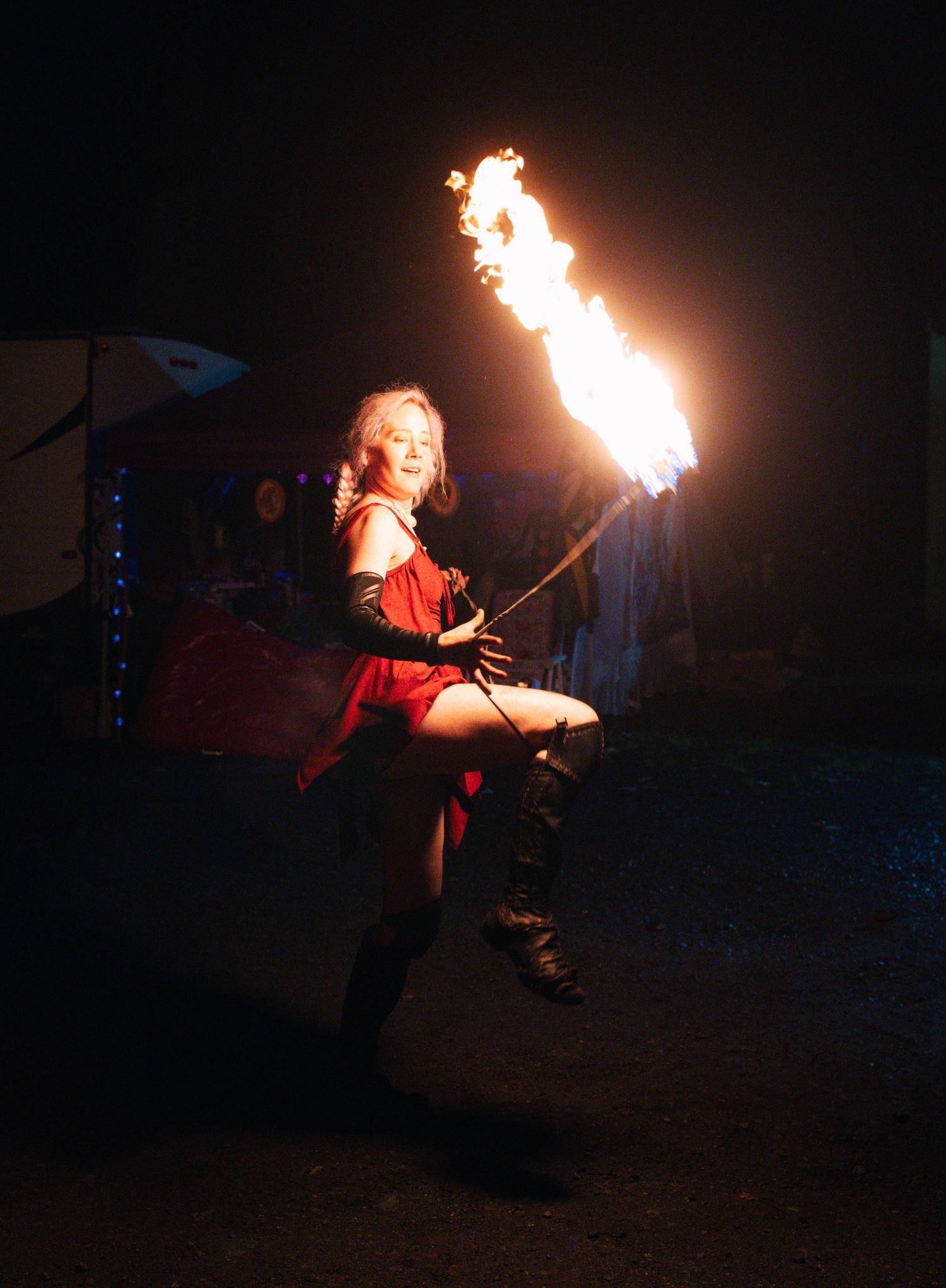 A female fire performer in a red costume twirls a flaming baton outdoors at night.