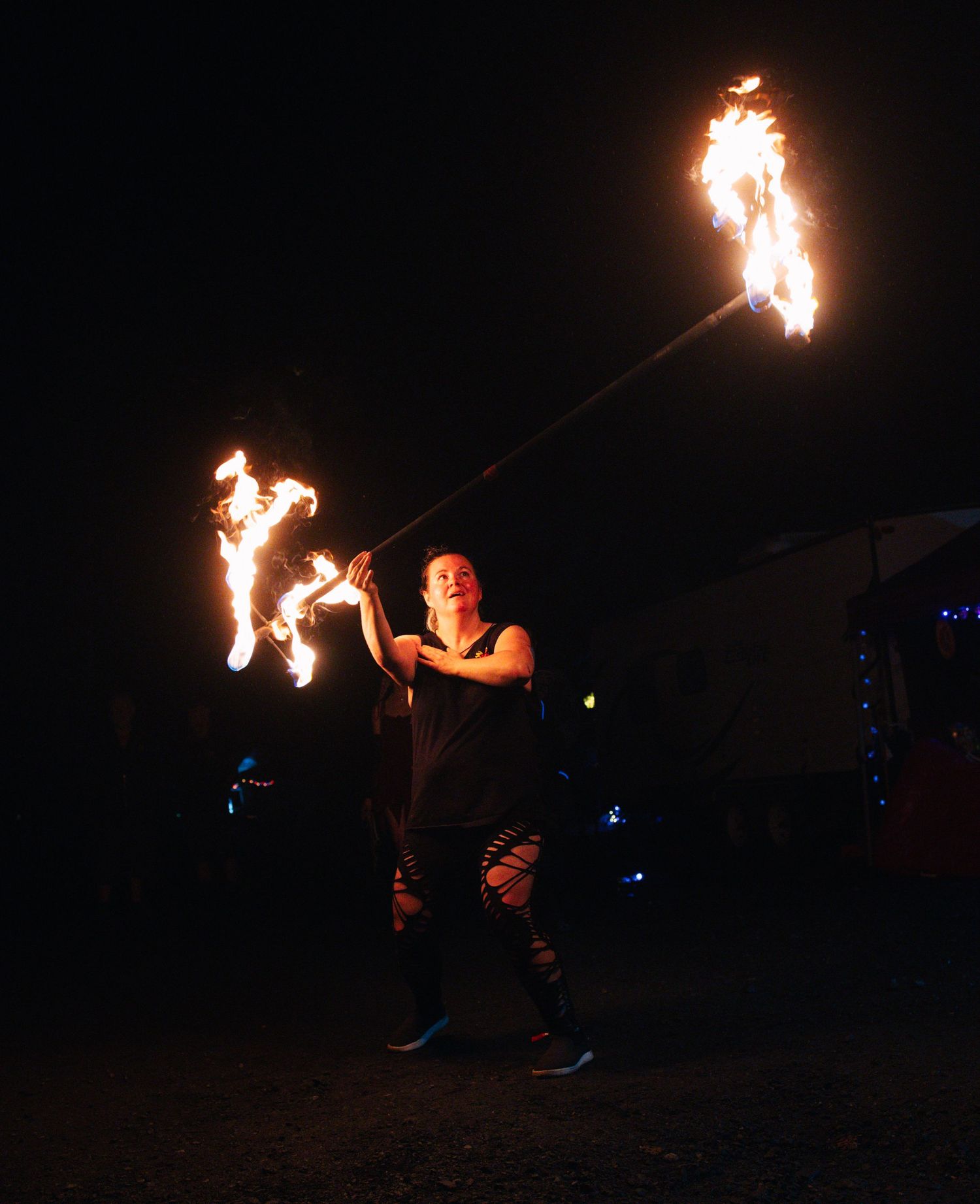 A fire performer twirls flaming batons dramatically against a dark night sky outdoors.
