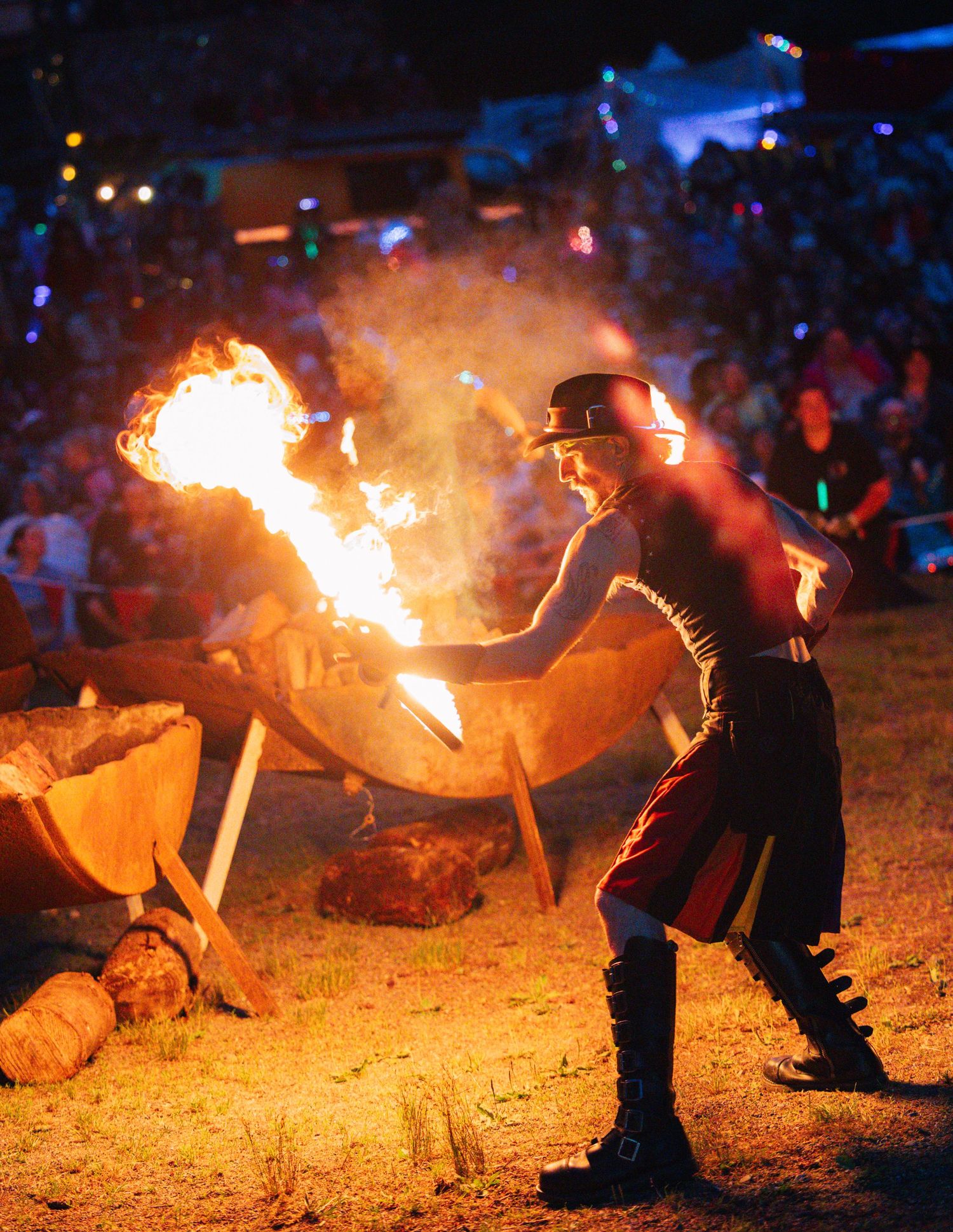 Fire performer breathing flames at a dramatic nighttime festival show, wearing costume with crowd watching in background.