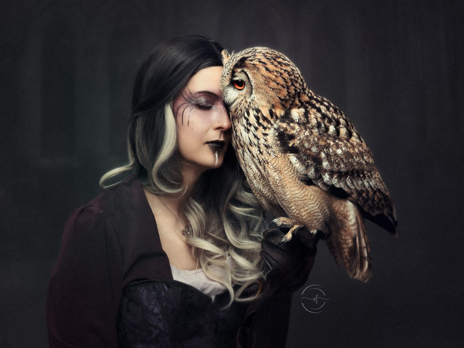 A woman with dark makeup and ombre hair poses with a large owl perched on her shoulder in a dramatic portrait.