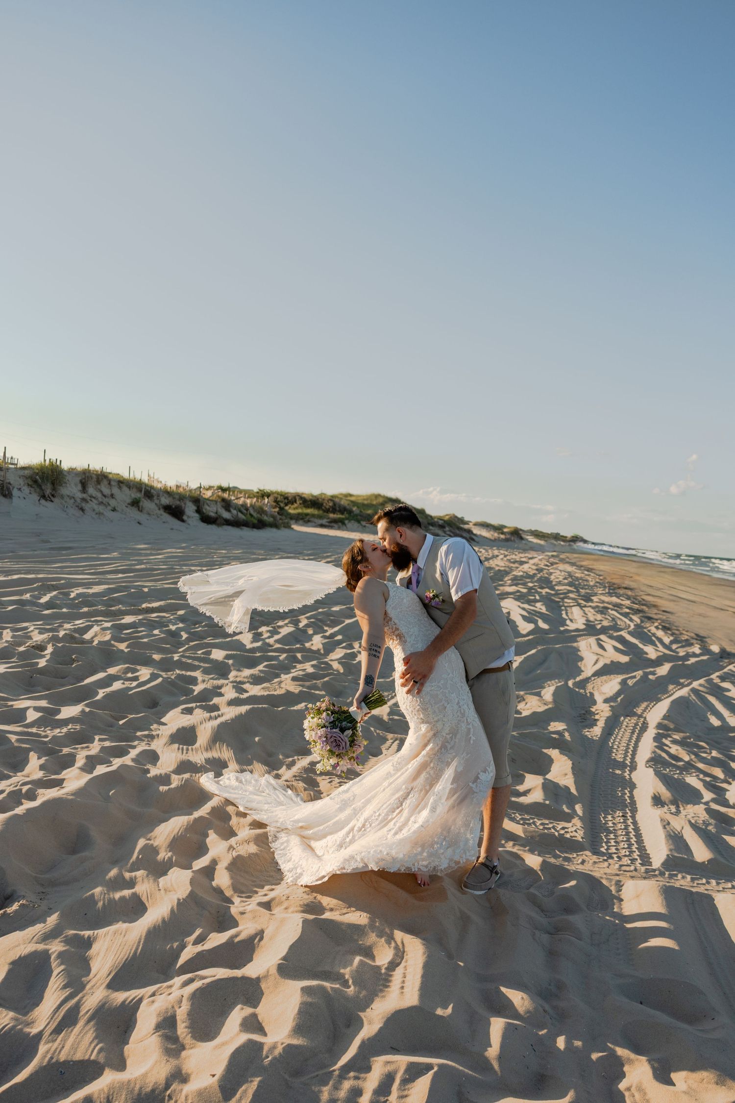 Bride and groom running joyfully on sandy beach at sunset, woman in flowing white dress, man in blue suit.