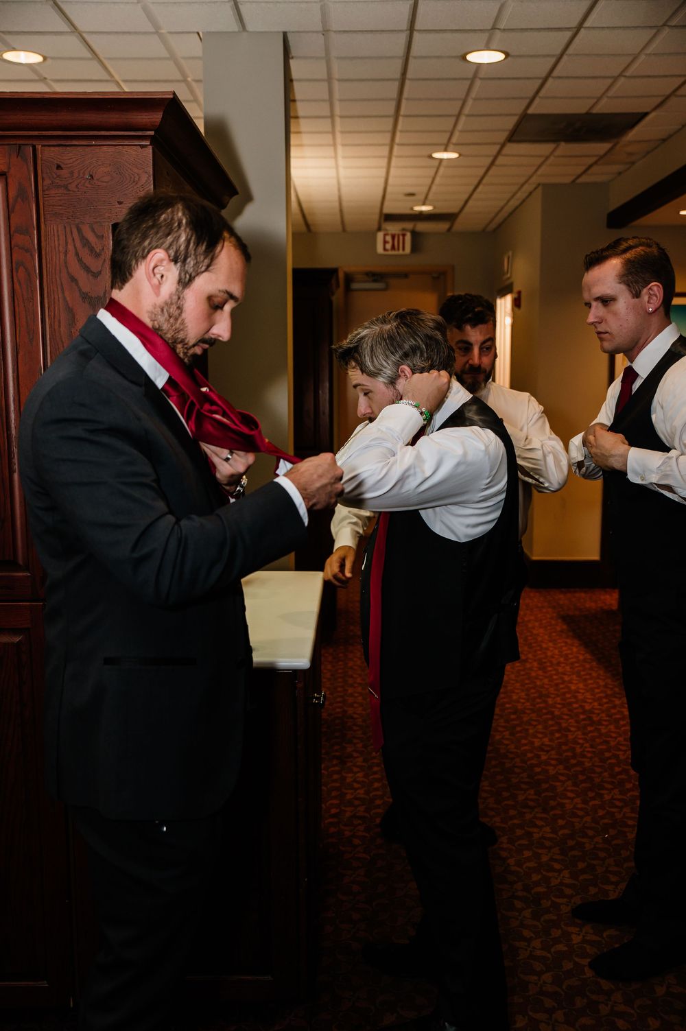 Groomsmen helping each other with red ties and boutonnieres in a hotel hallway before a wedding ceremony.