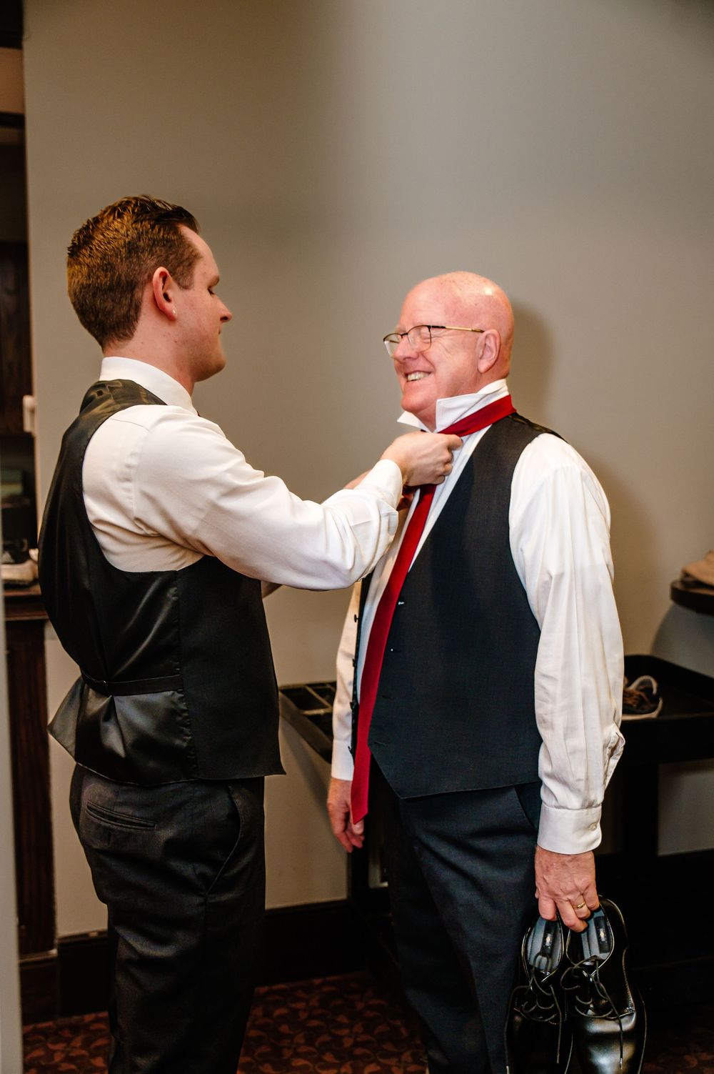 Groom helping older man adjust red tie while both wear black vests before wedding ceremony.