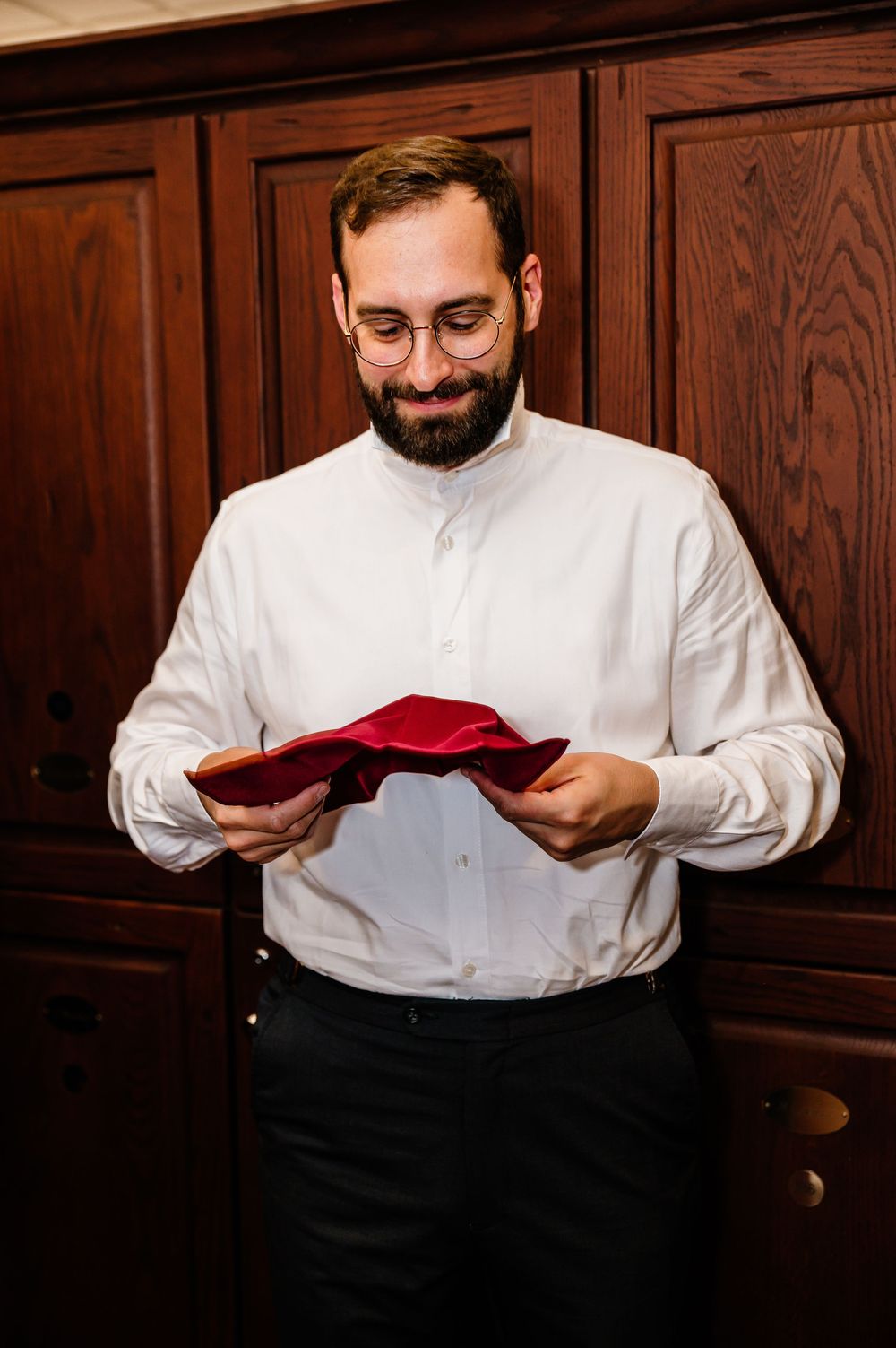 Bearded man with glasses in white shirt holding a red pocket square, preparing for a formal event.