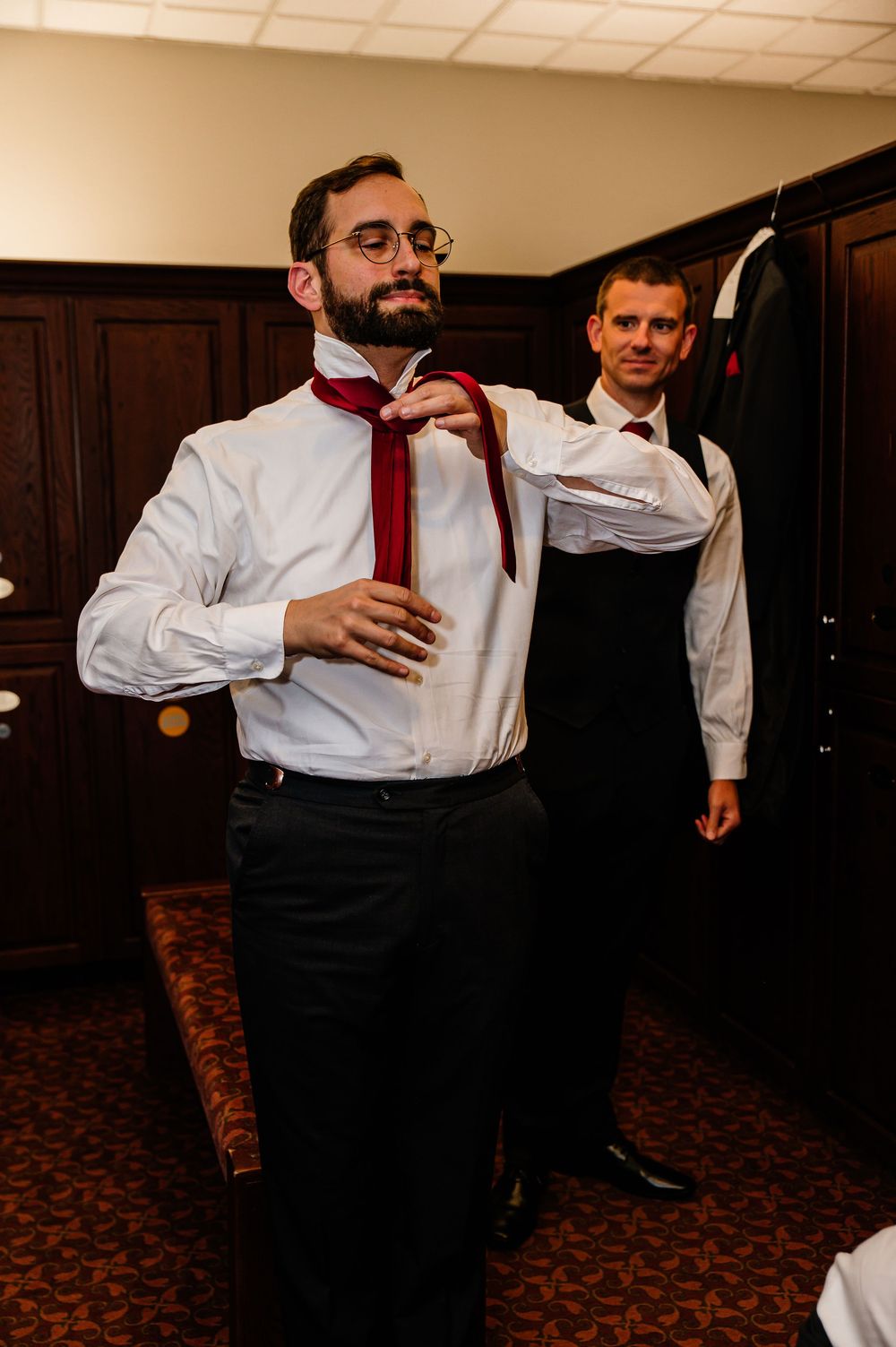Groom adjusting red tie with help from groomsman in vest before wedding ceremony in elegant room.