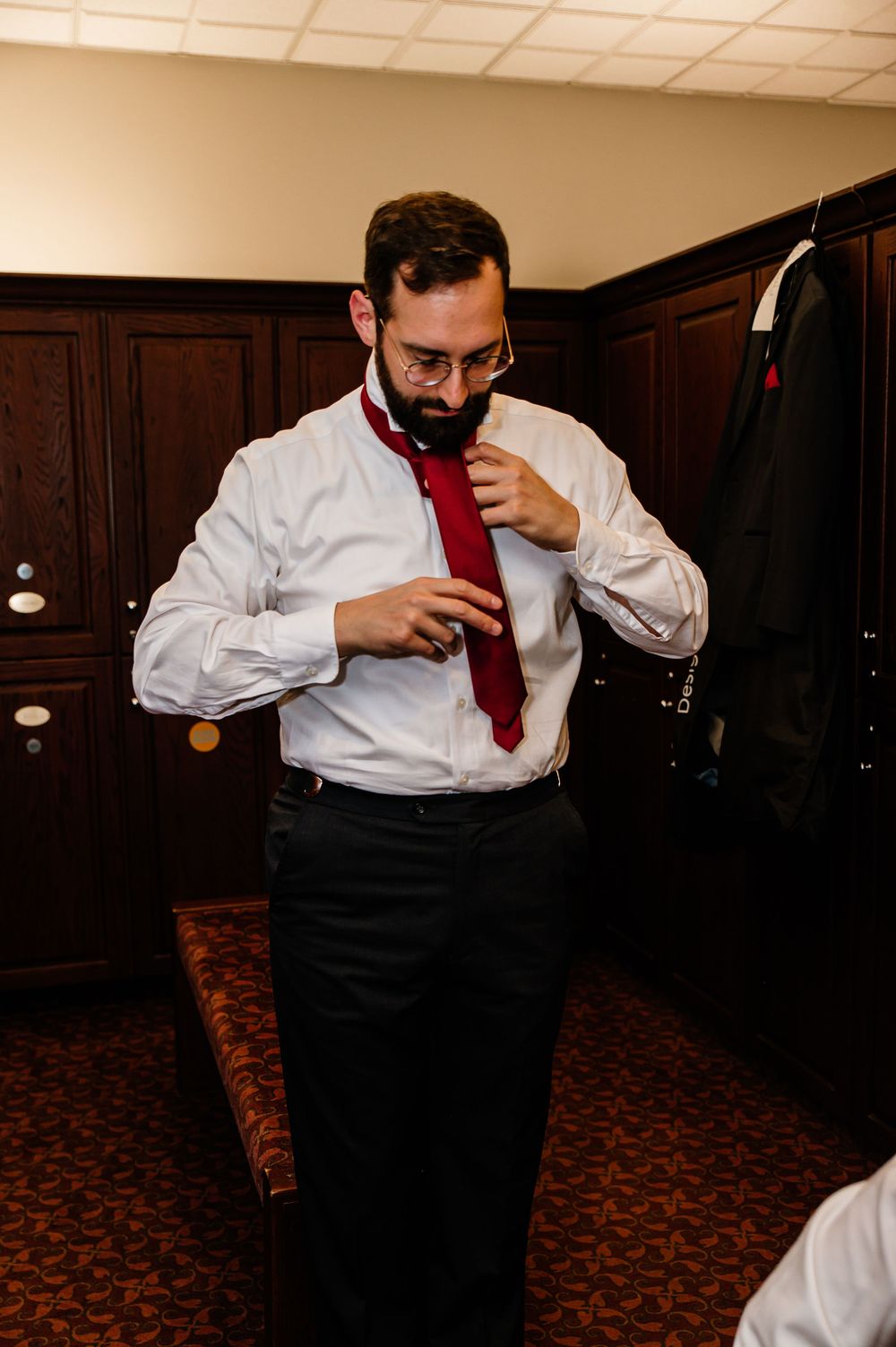 A groom in a white dress shirt adjusts his red tie while getting ready in a wood-paneled dressing room.