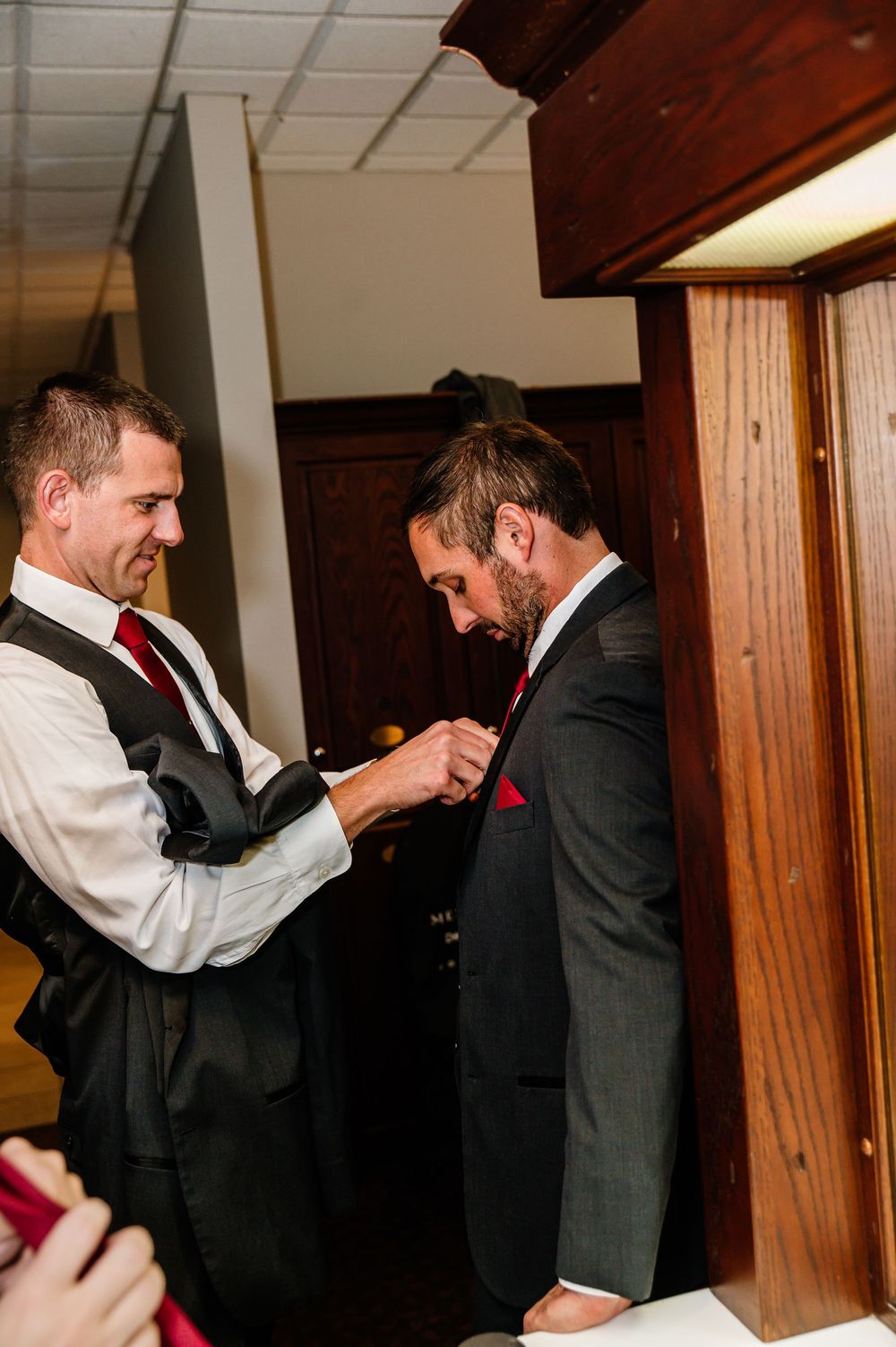 Two groomsmen in dark suits with red ties help each other with boutonnieres before a wedding ceremony.