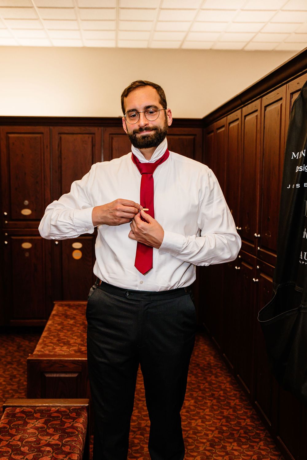 A bearded man in a white shirt and red tie adjusts his shirt buttons in a wood-paneled locker room.
